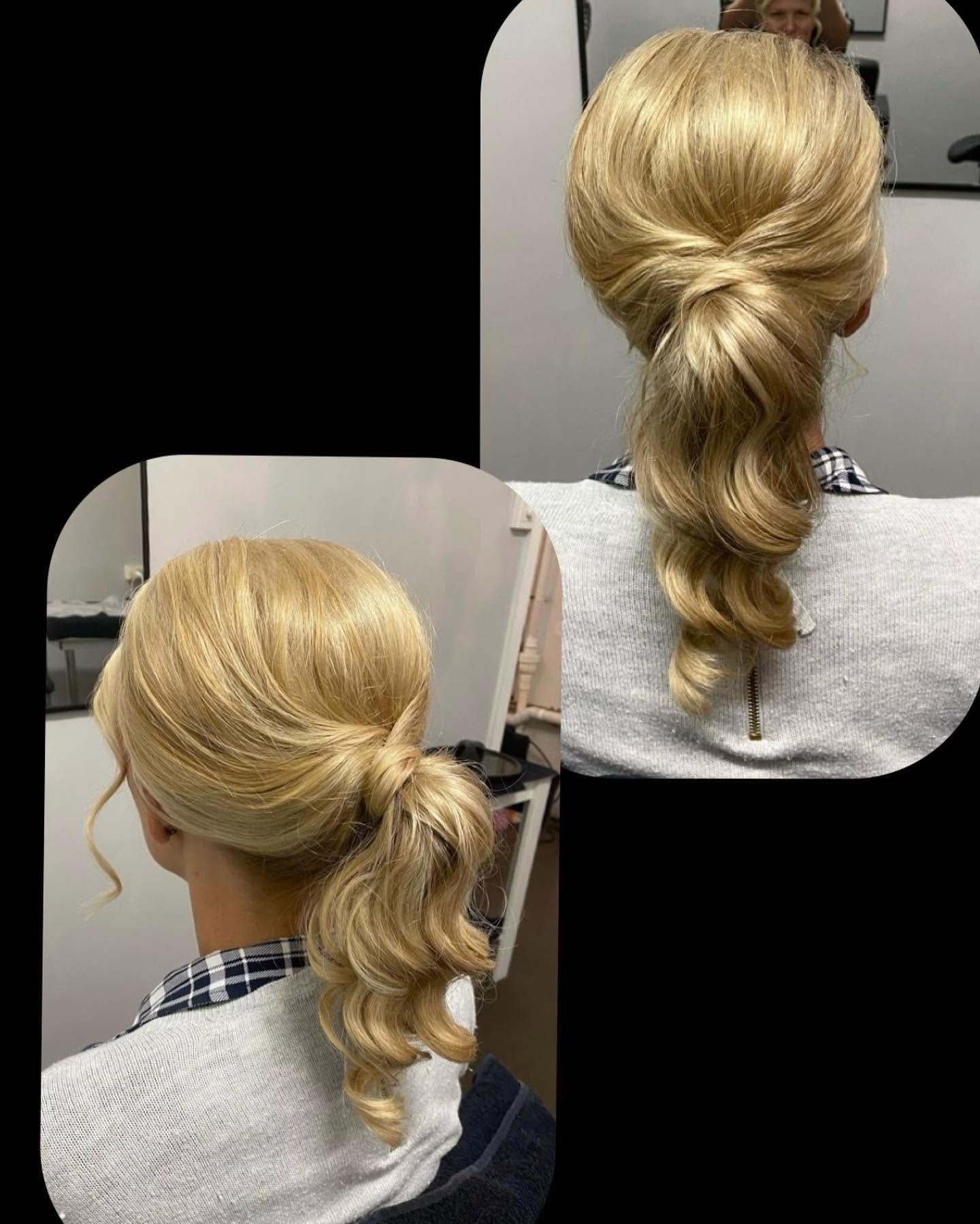 A woman with blonde hair styled in a twisted low bun with a loose curl free hanging. The woman is sitting in a salon chair wearing a checkered shirt and a gray cardigan, with a mirror reflecting her and the salon setting.