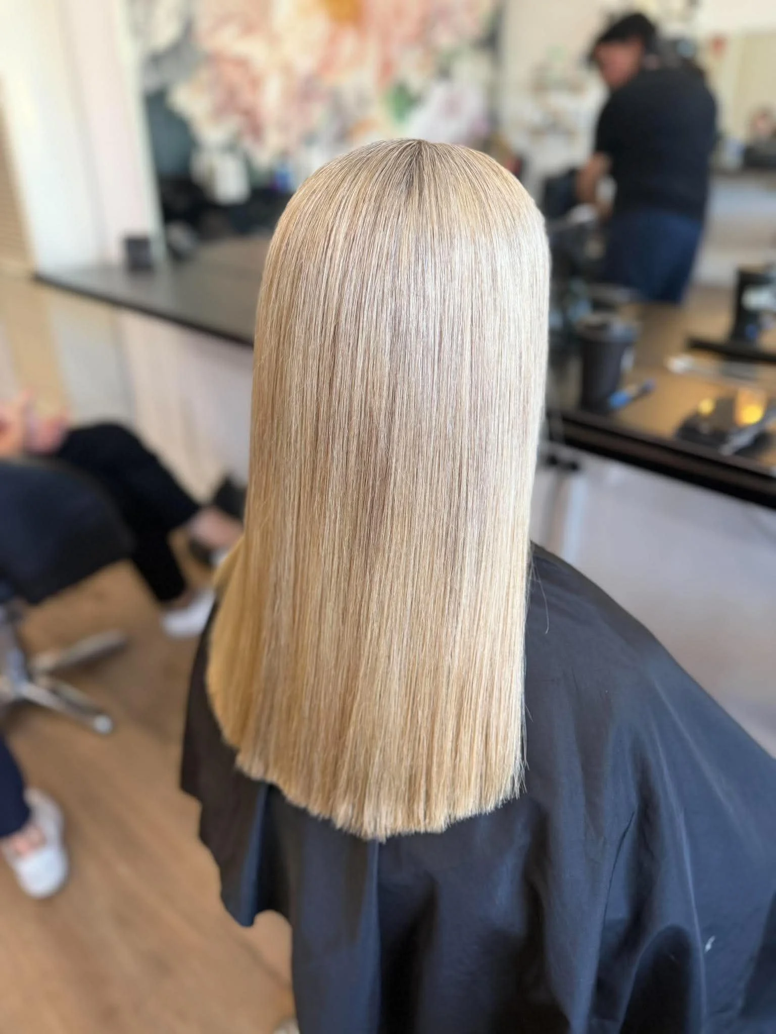 Close-up of a person with long, straight, blonde hair sitting in a salon, with a stylist working in the background.