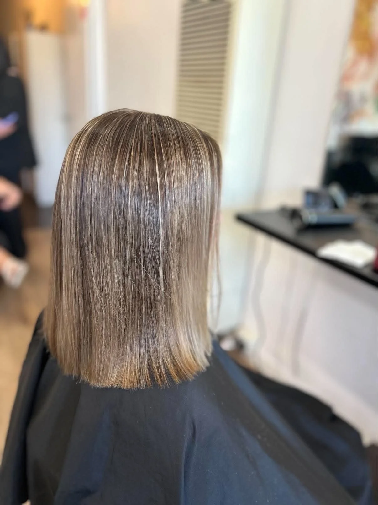 Back view of a woman with straight, shoulder-length, light brown hair with highlights, inside a salon.