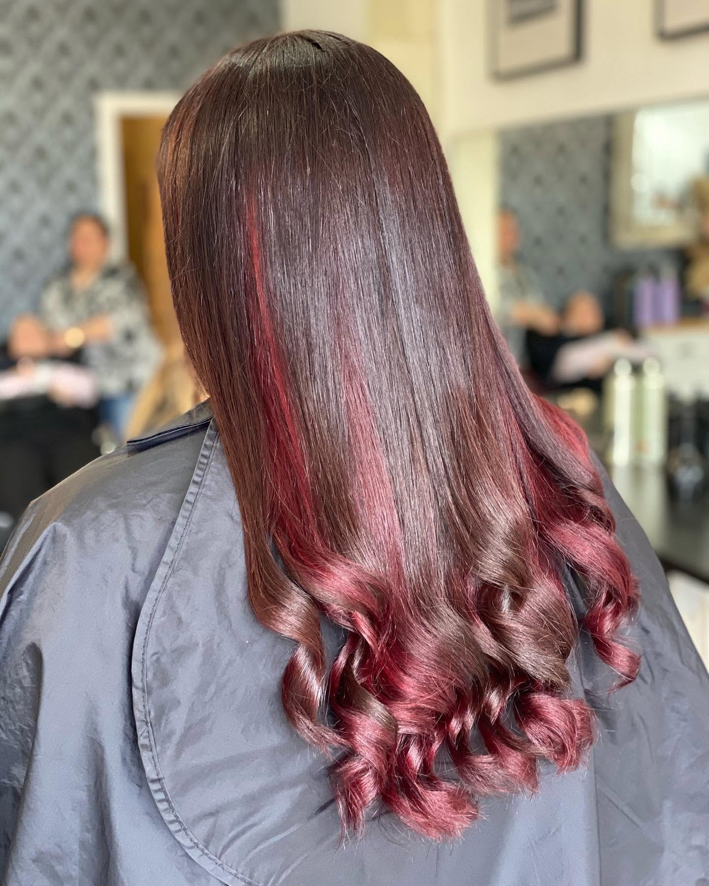 Back view of a person with shiny, straight brown hair that transitions into red, styled in loose curls, in a salon setting.