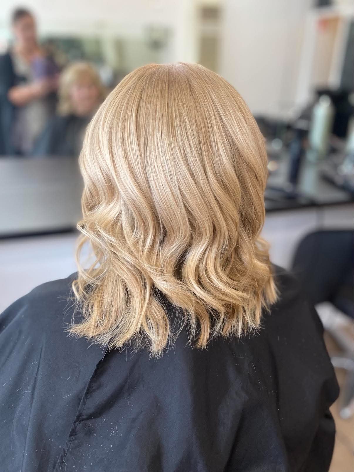 Back of a person's head with shoulder-length, wavy blonde hair, sitting in a salon with a mirror and hairstyling tools visible.