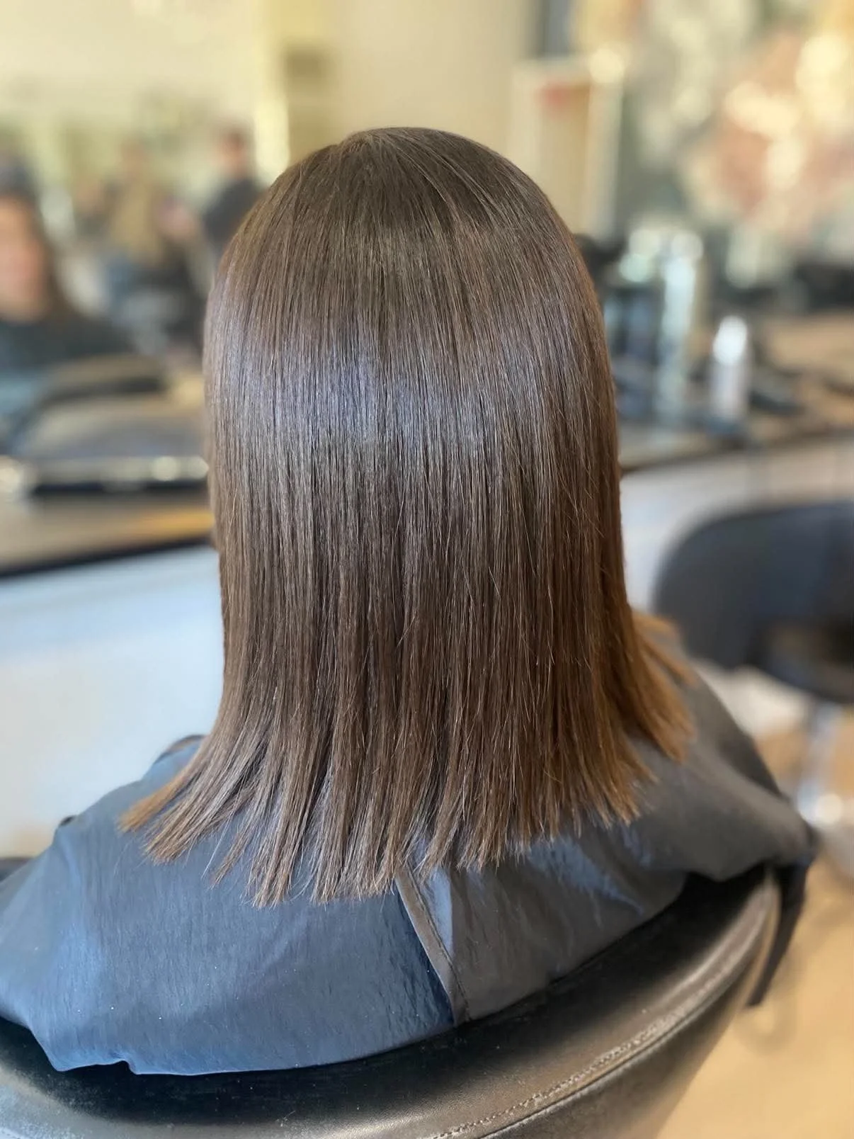 Back of a person's head with straight, shiny brown hair in a salon setting.