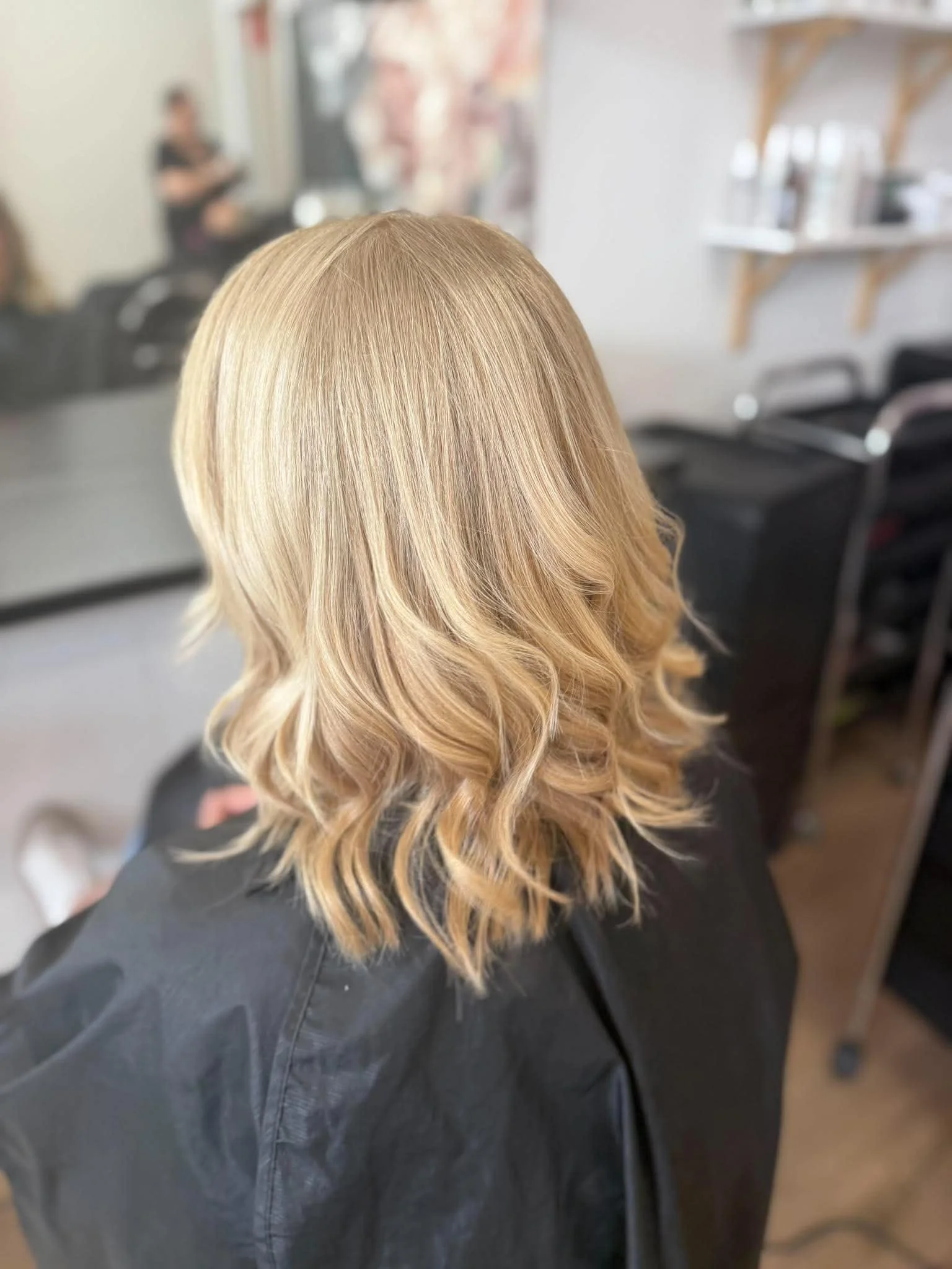 Blonde woman with wavy hair sitting in a salon chair, viewed from the back. Salon with chairs and a hairstylist in the background.
