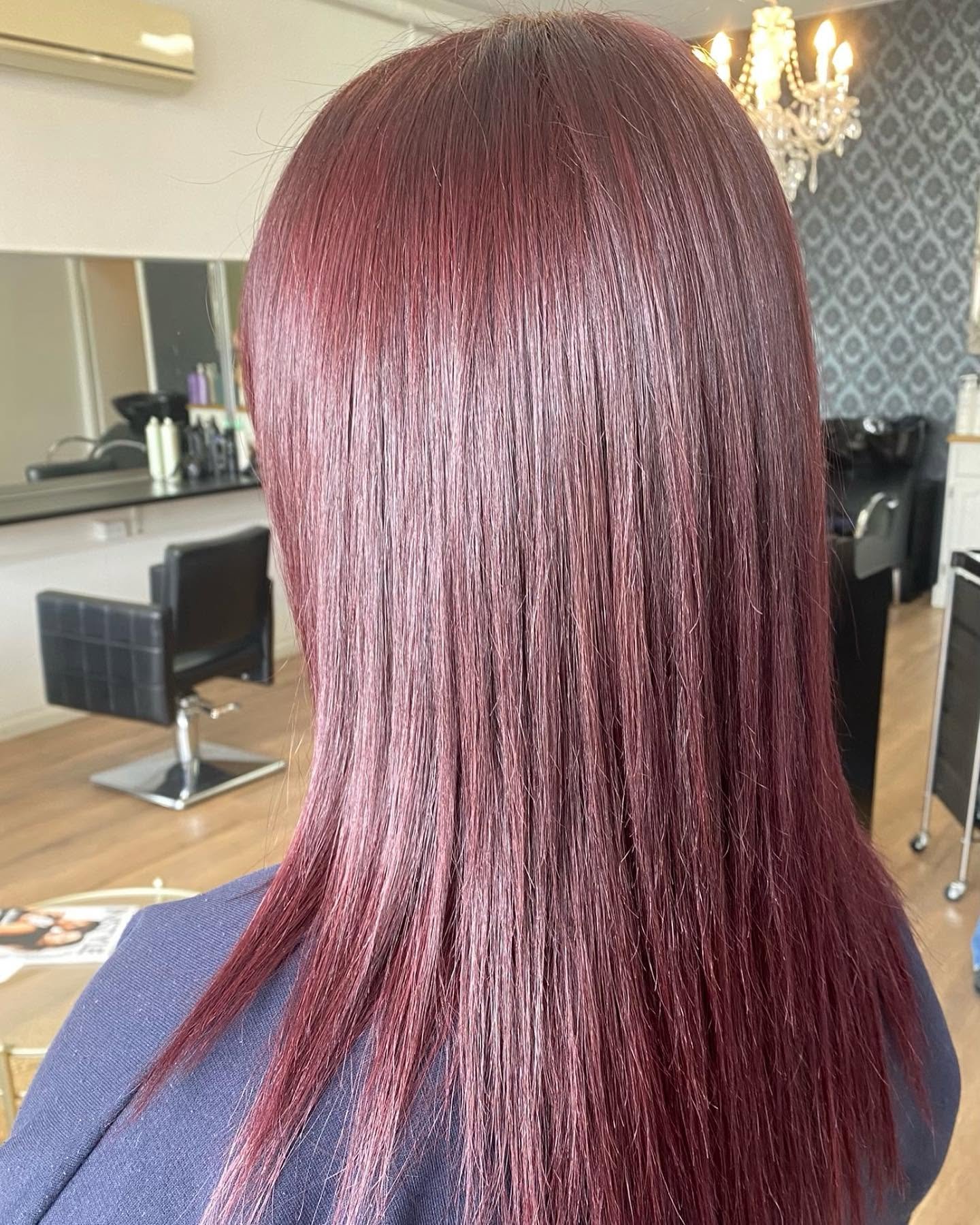 Back view of a woman with long, straight, burgundy-colored hair in a salon setting, with black salon chairs, a mirror, and a chandelier visible in the background.