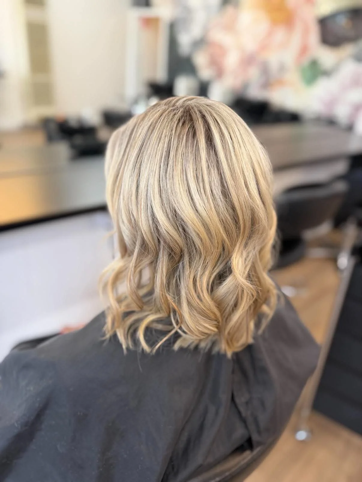Back view of a person with shoulder-length blonde wavy hair in a salon setting.