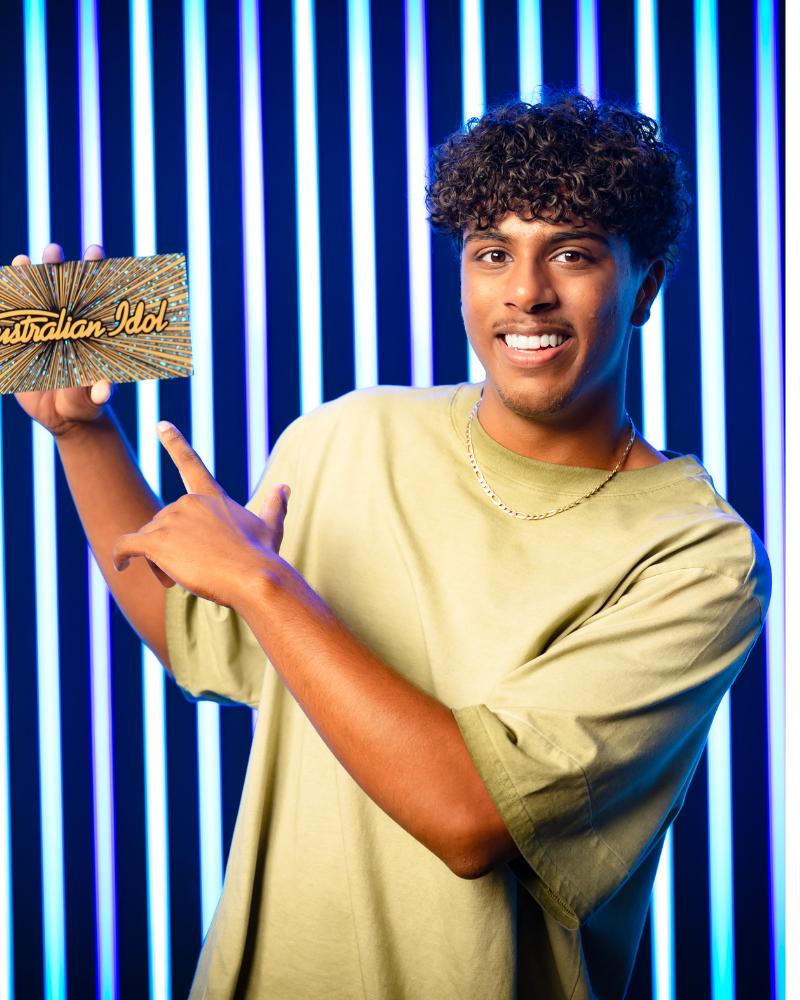 Young man with curly hair smiling and pointing at a sign that reads "Australian Idol" against a background of vertical blue neon lights.