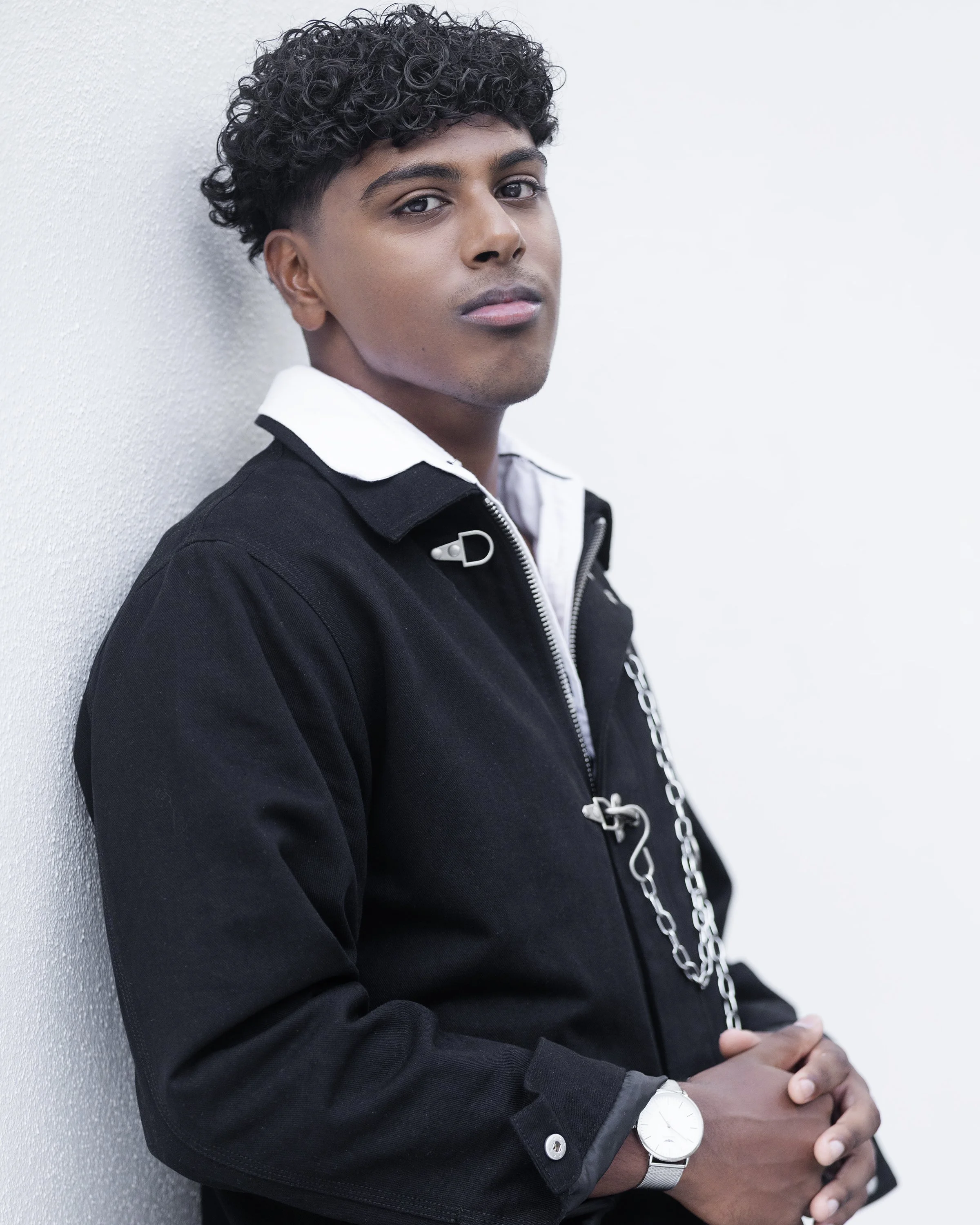 A young man with curly black hair leaning against a white wall, wearing a black jacket, white shirt, silver watch, and displaying a neutral expression.