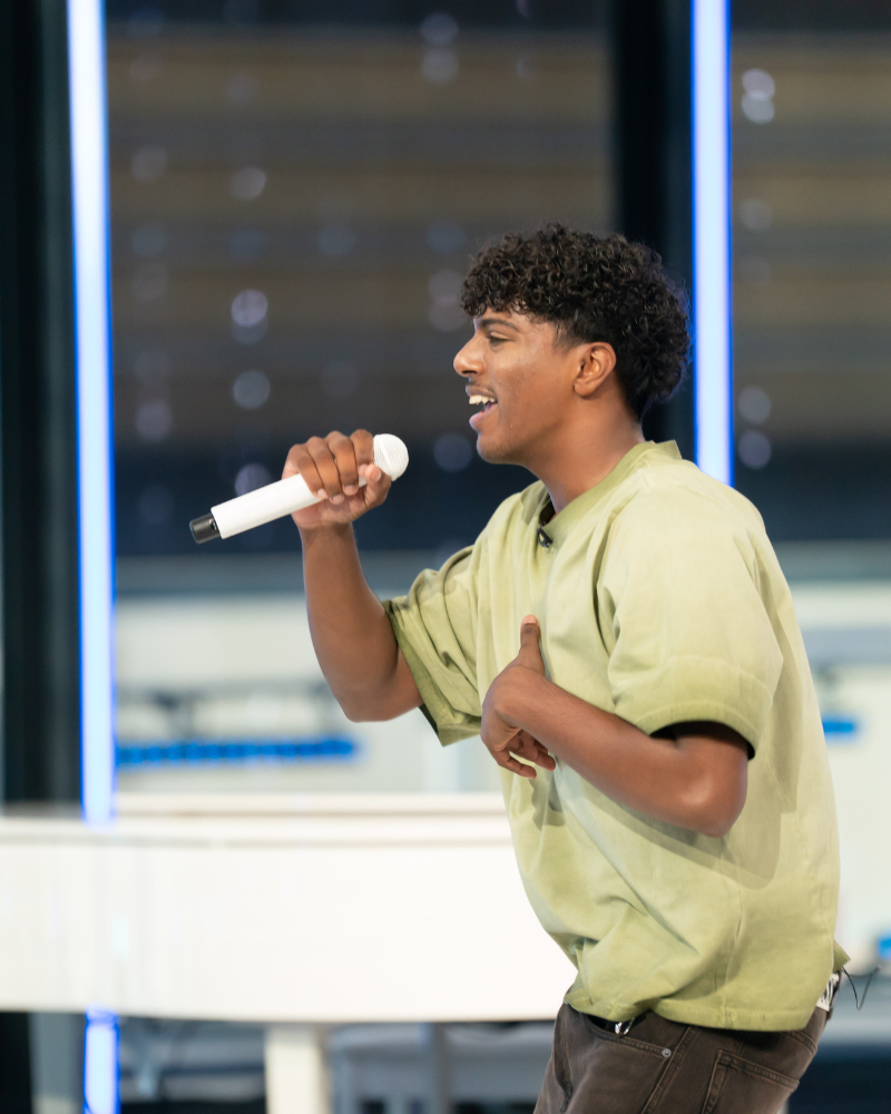 A young man with curly hair smiling and singing into a microphone.