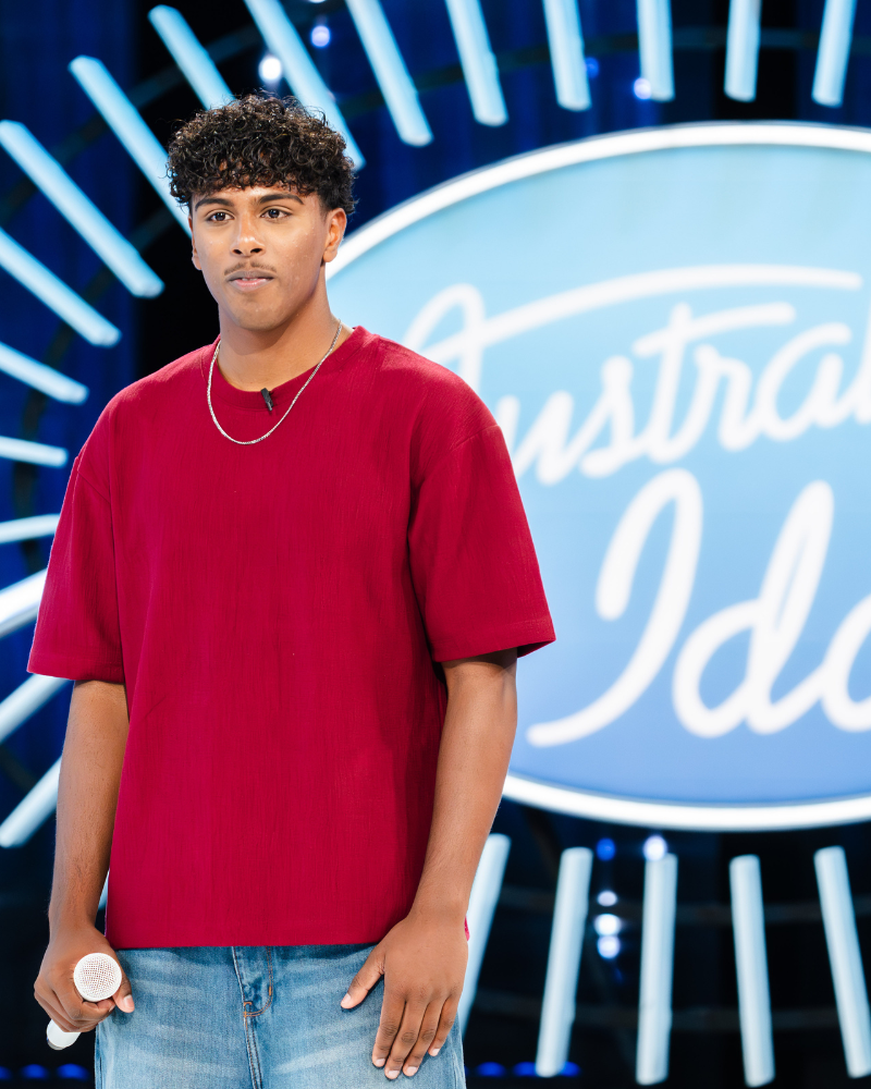 Young man with curly hair standing in front of an American Idol logo on stage, holding a microphone, wearing a red t-shirt and jeans.