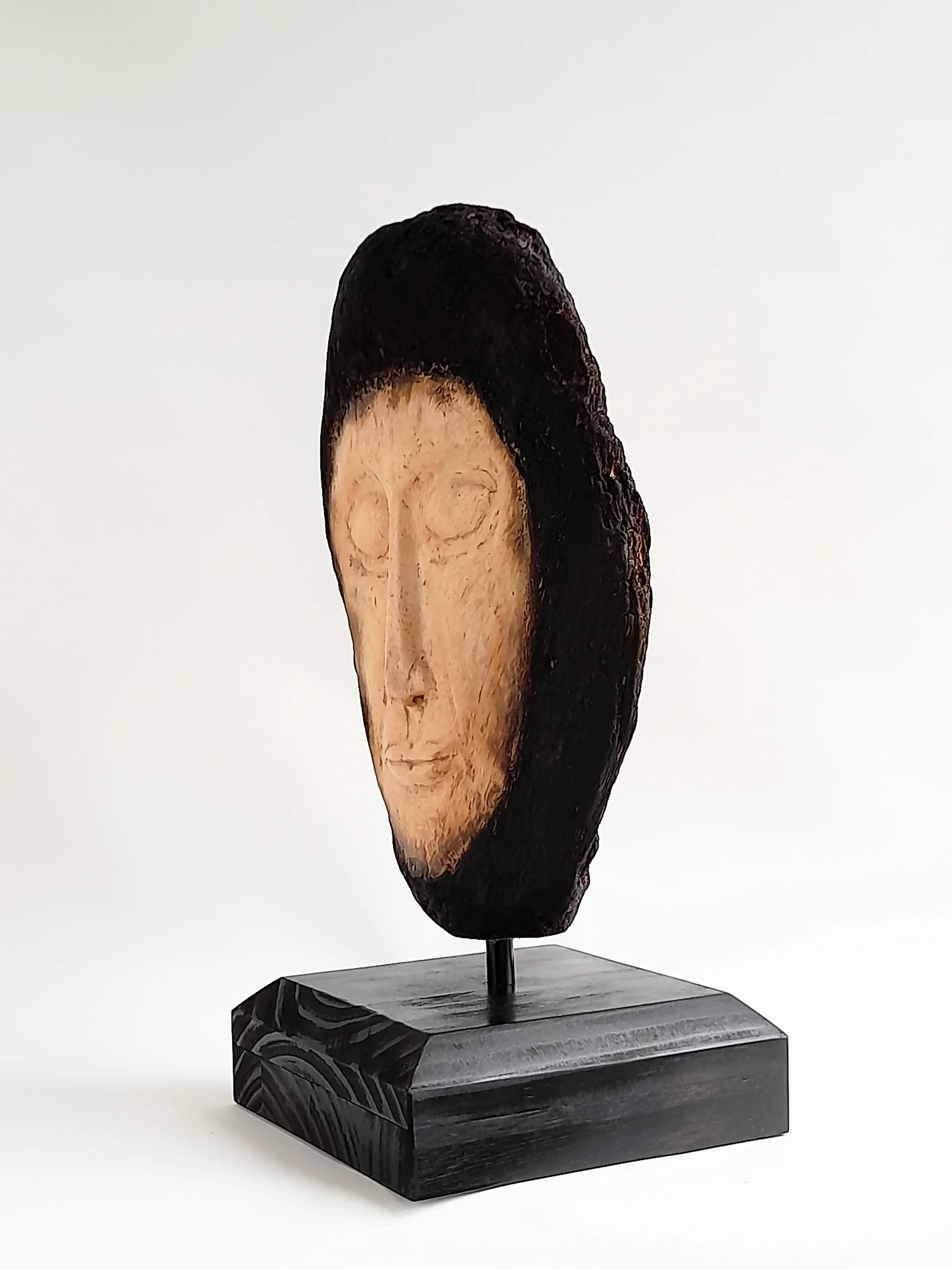 Hand-carved pale wooden face sculpture framed by charred dark timber on a black pedestal titled Beyond the Char.