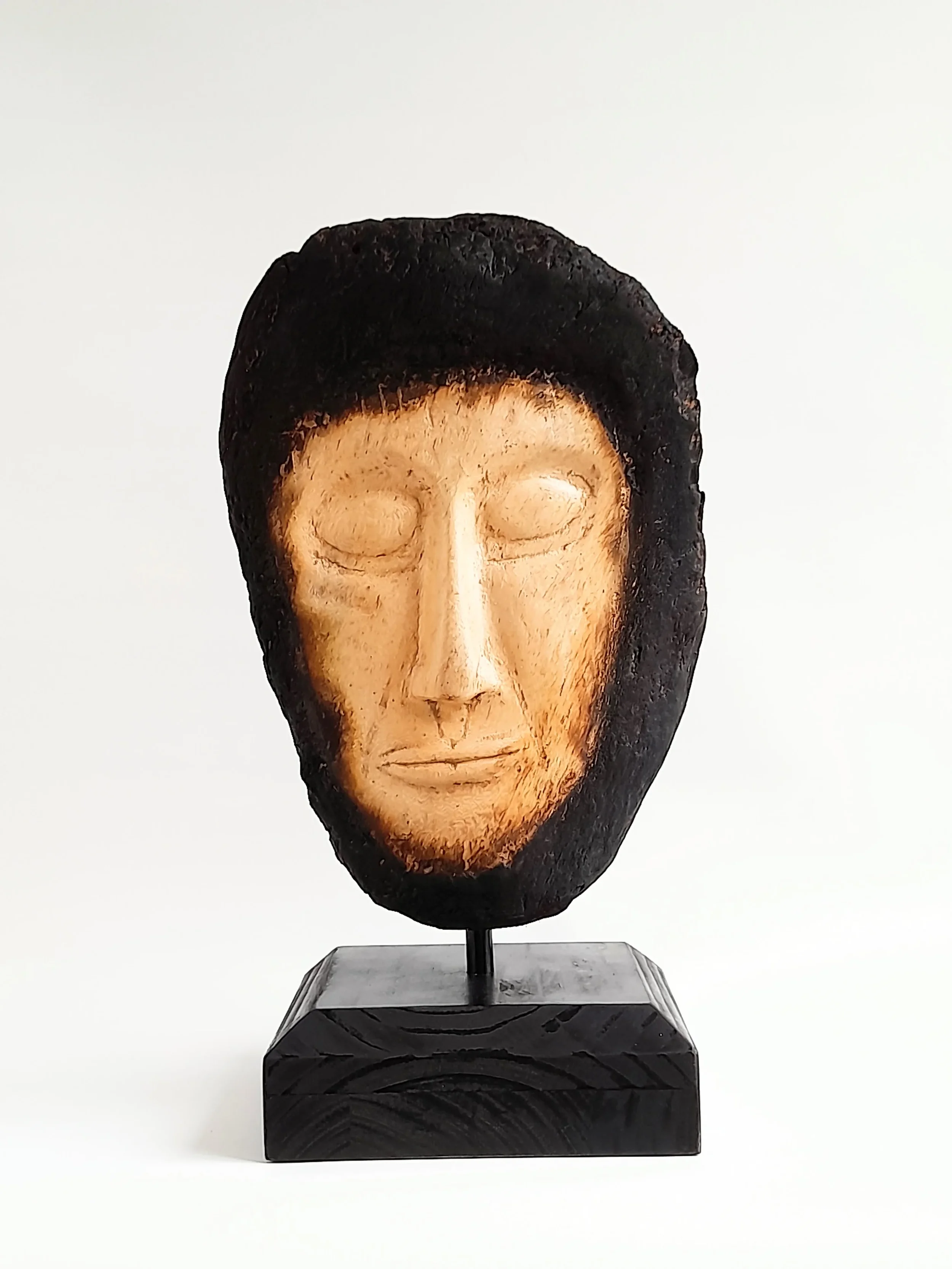 Hand-carved pale wooden face sculpture framed by charred dark timber on a black pedestal titled Beyond the Char.