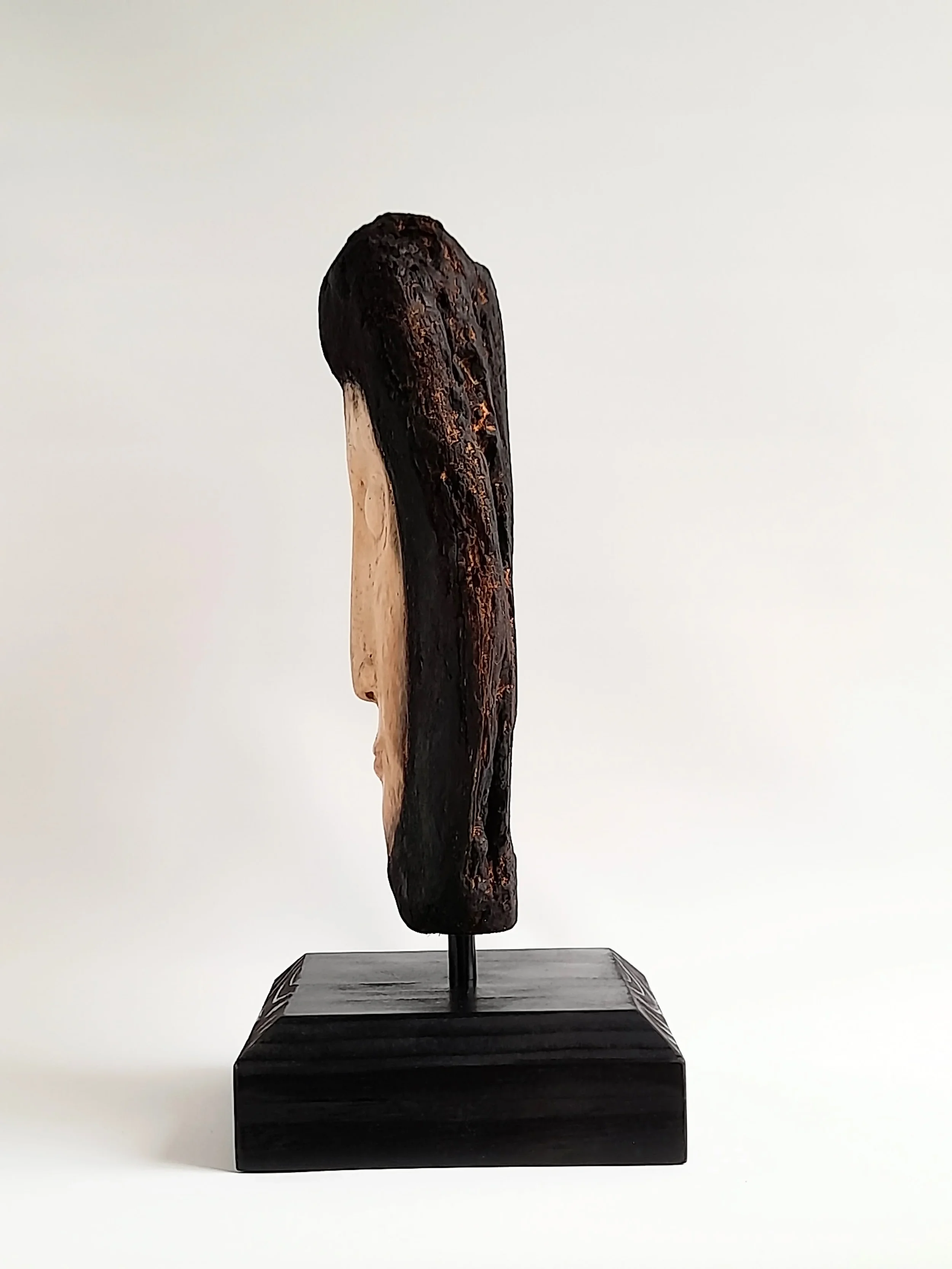 Hand-carved pale wooden face sculpture framed by charred dark timber side view titled Beyond the Char.