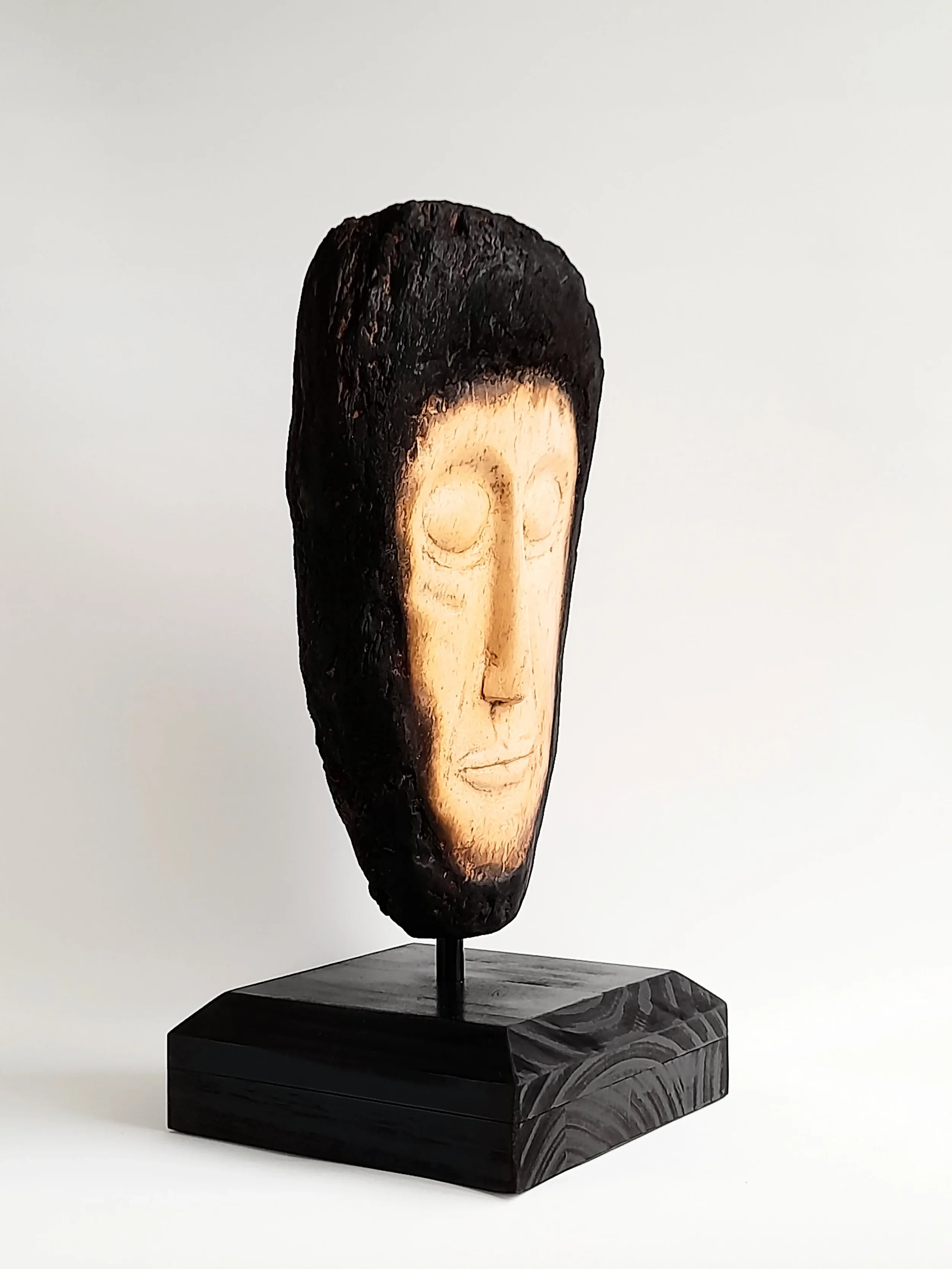 Hand-carved pale wooden face sculpture framed by charred dark timber on a black pedestal titled Beyond the Char.