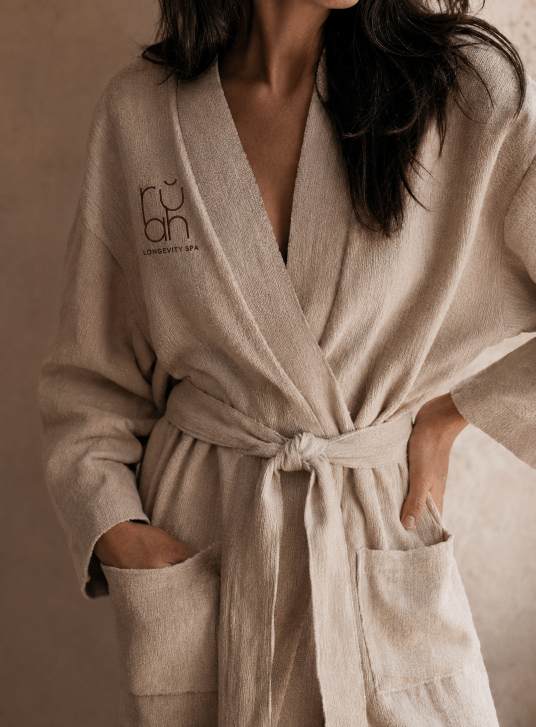 Woman wearing a beige bathrobe with a logo and text on the left side that says "LÖVE Longevity Spa," standing with hands in pockets.