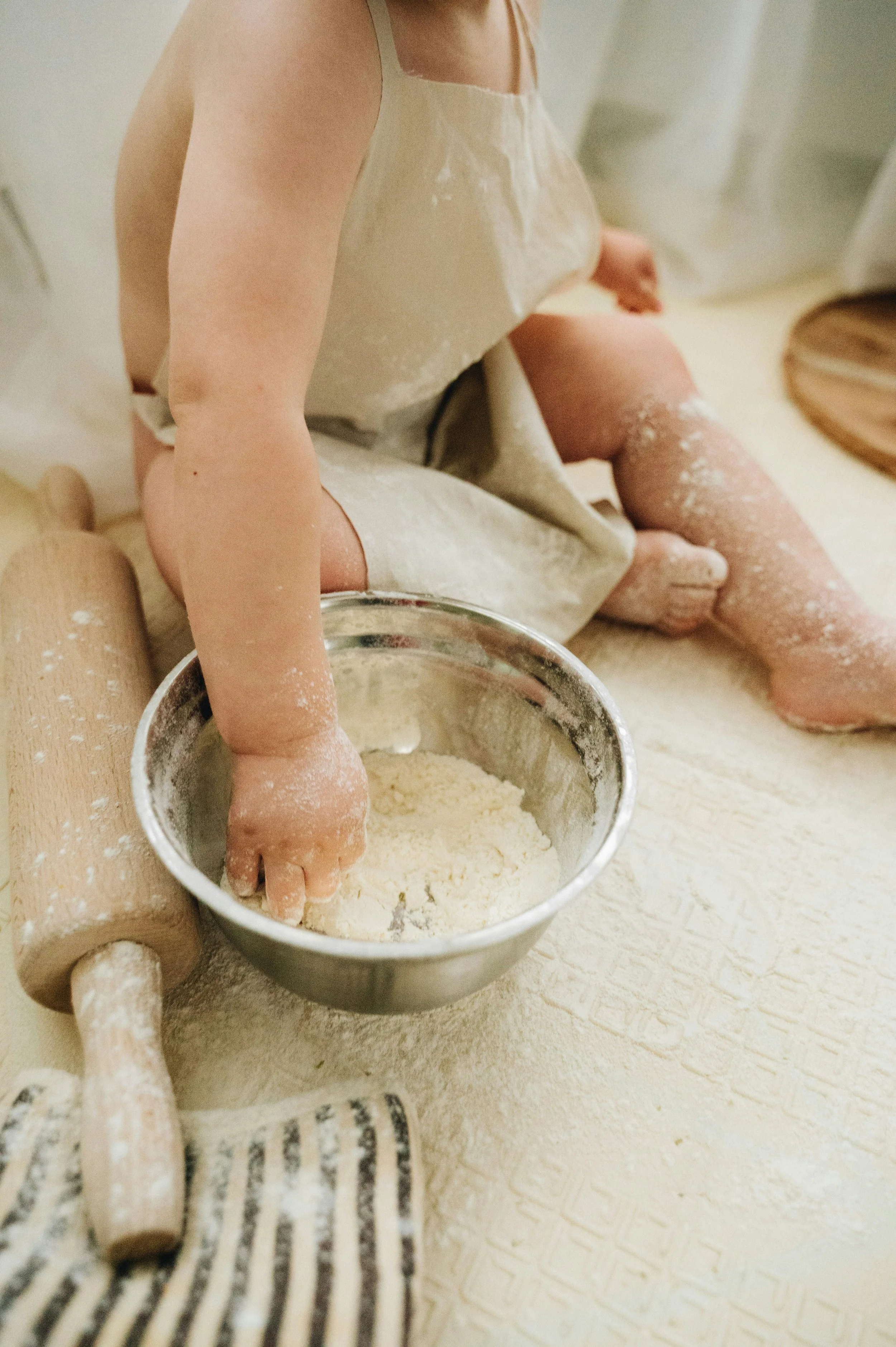 Child in a beige apron playing with flour and measuring cups on a kitchen counter.