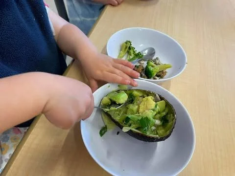 A child's hands reaching for a bowl of guacamole on a wooden table, with a bowl of broccoli nearby.