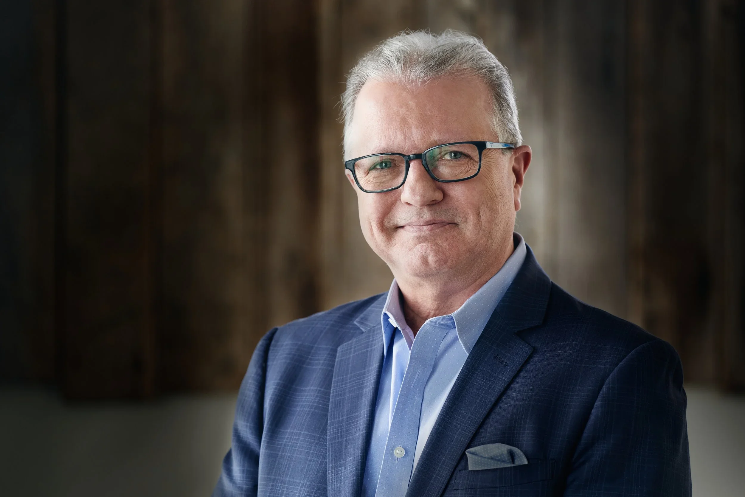 A middle-aged man with gray hair, wearing black-rimmed glasses, dressed in a blue checked suit and light blue shirt, smiling confidently against a blurred background.