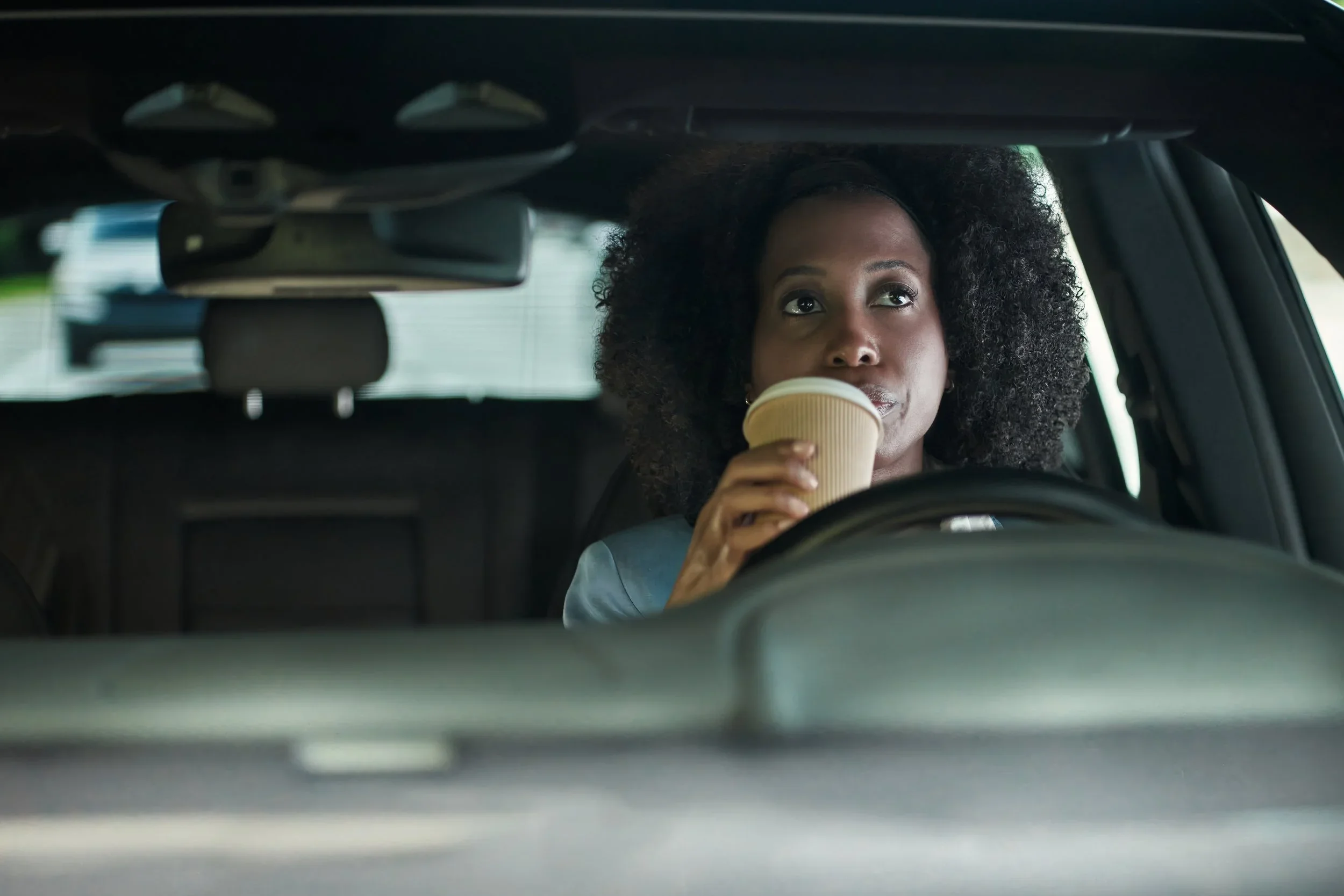 Woman with curly hair drinking coffee from a paper cup while sitting in the driver's seat of a car.