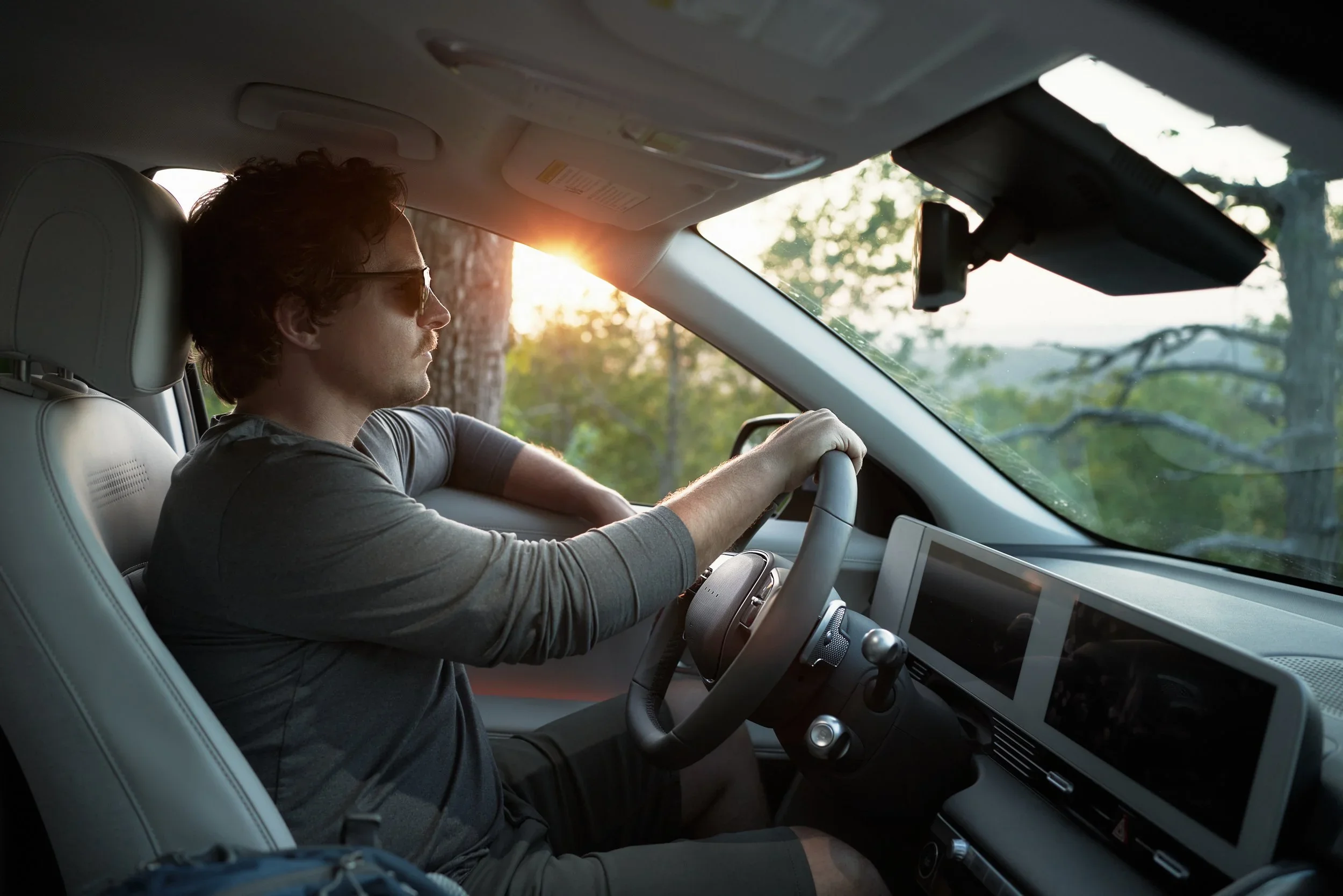 A man with curly hair and sunglasses driving a car during sunset, surrounded by trees and nature.