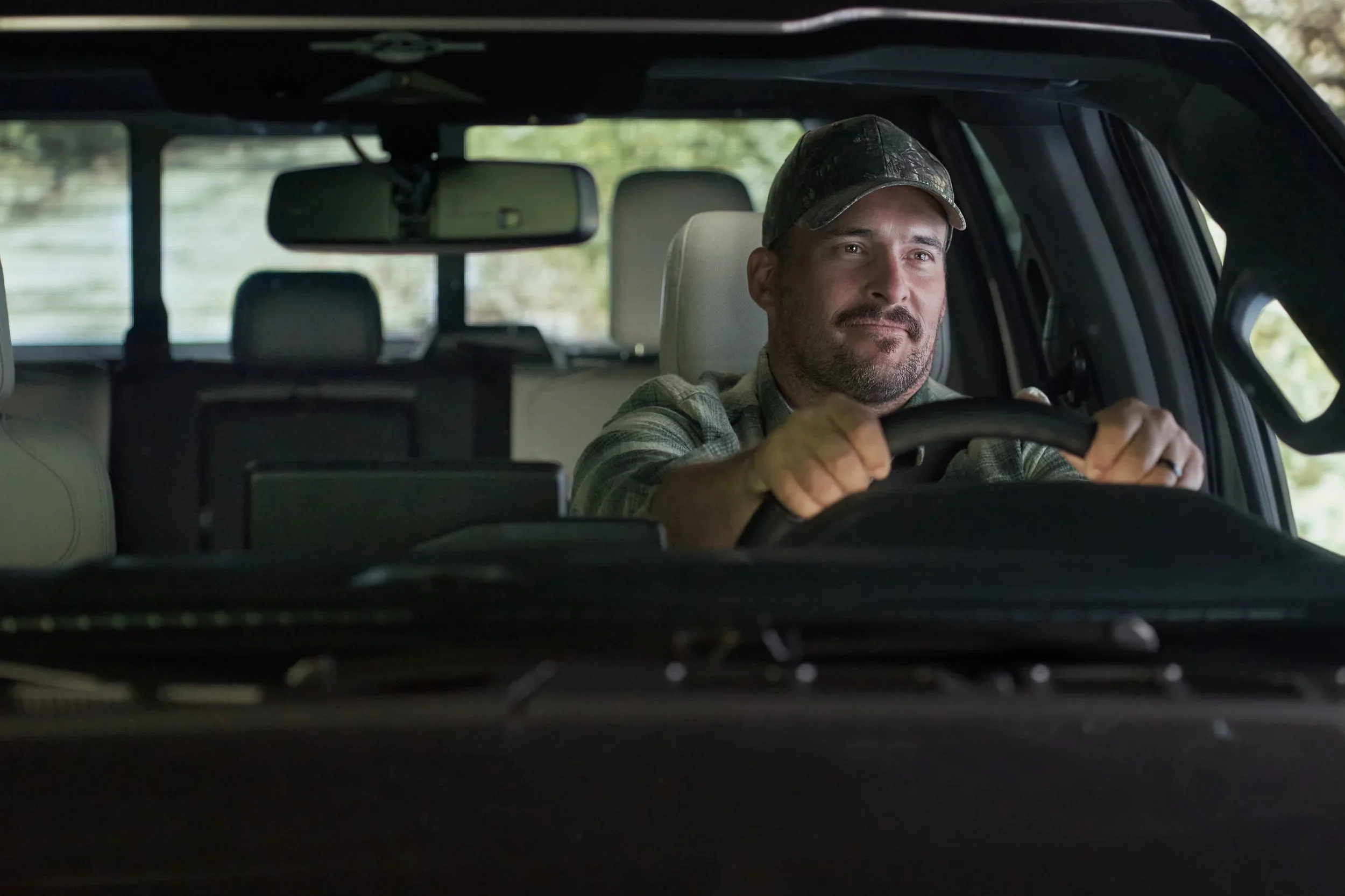 A man wearing a camouflage cap and plaid shirt driving a vehicle, with trees visible outside the window.