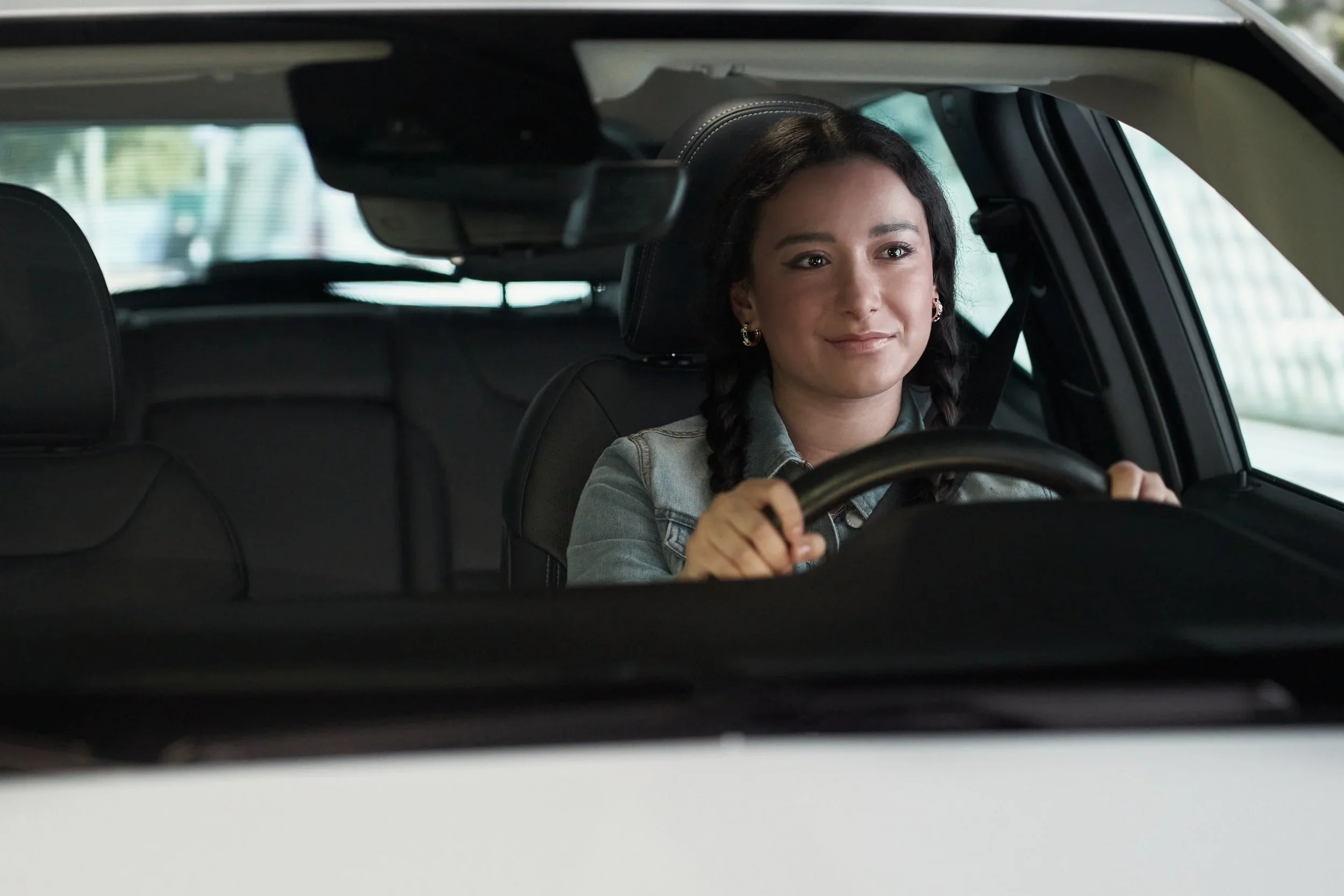 Young woman with braided hair and earrings driving a car, viewed through the rearview mirror.