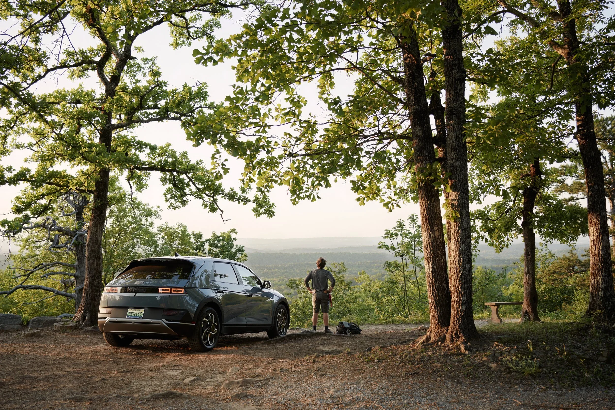 A person standing on a trail near a black SUV, overlooking a scenic view of trees and a distant landscape at sunset, in a wooded area with tall trees and a parking area.