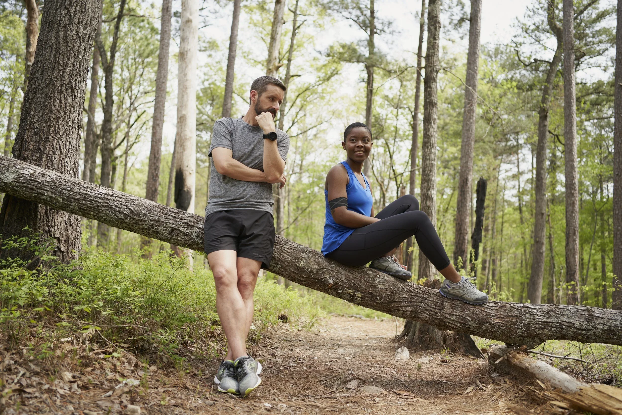 A man and woman enjoy a hike in a dense green forest, with the woman sitting on a fallen tree and the man standing nearby, both looking relaxed.