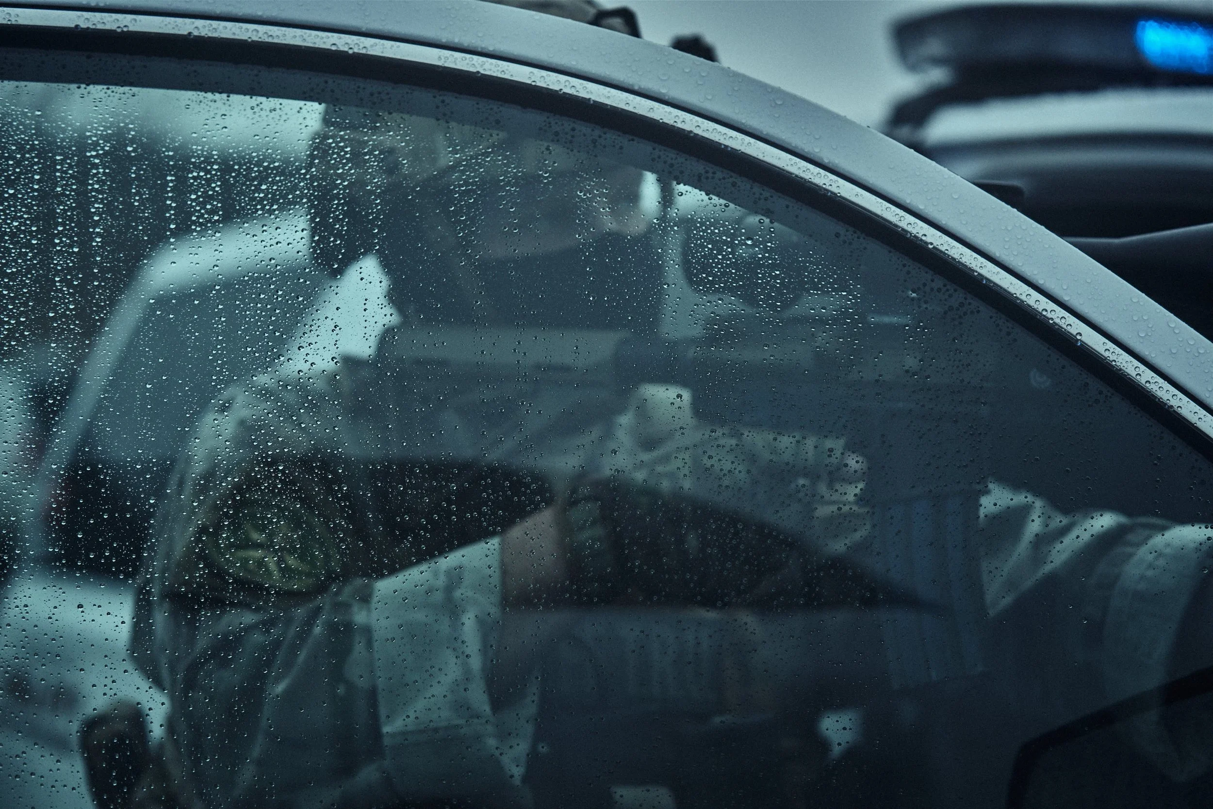Inside a vehicle during rain, raindrops on the windshield, with a blurred view of a person in the driver's seat.