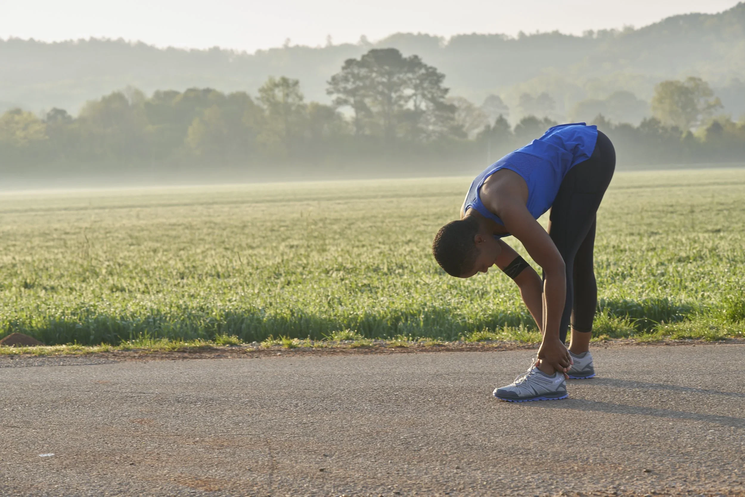 A person stretching and tying their shoe on a running track in a park with trees in the background.