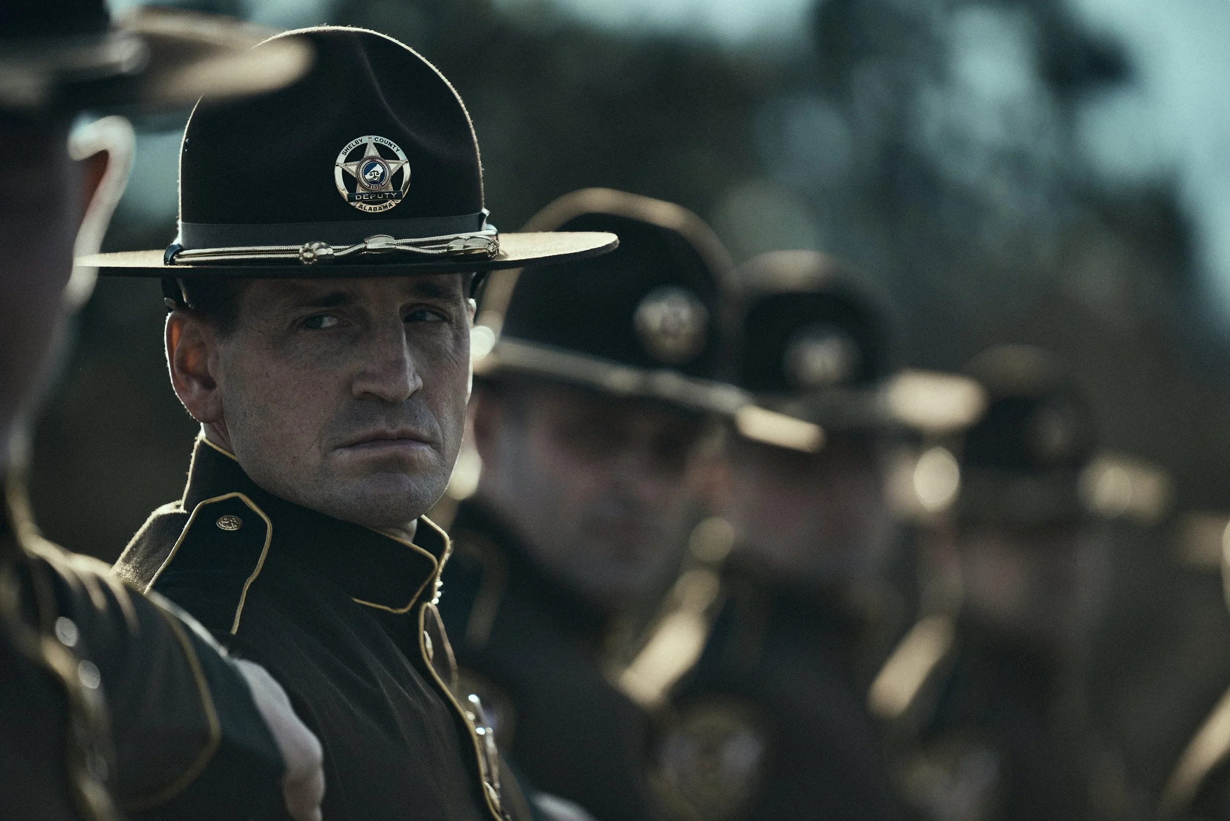 Close-up of a line of uniformed police officers, with focus on a police officer wearing a hat with a sheriff's badge, in a parade or formal event setting.