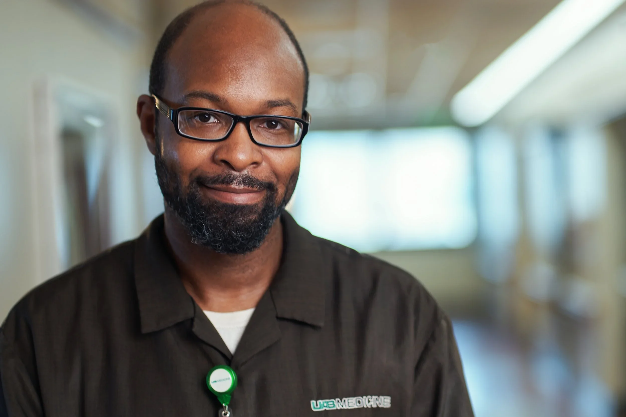 A man with glasses and a beard wearing a black shirt with a green emergency contact badge around his neck, standing in a hallway with bright natural light.