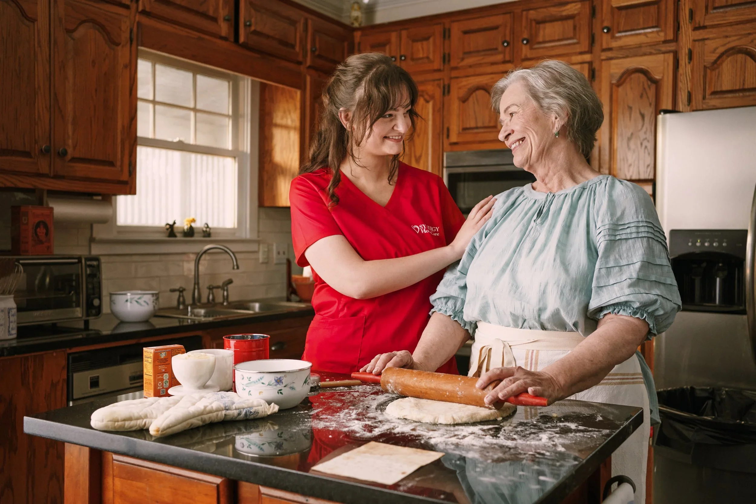 A young woman wearing a red shirt helping an elderly woman in a kitchen. They are smiling at each other while baking. The kitchen has wooden cabinets, a window, and baking ingredients on the counter.