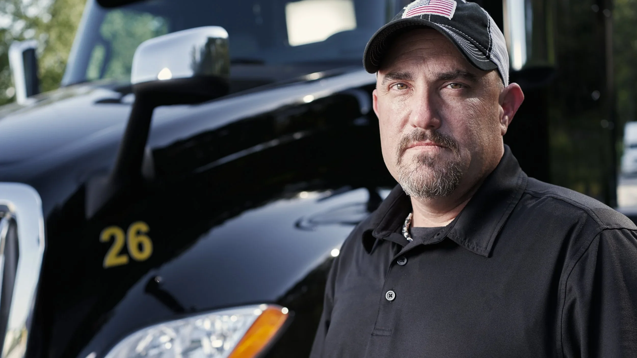 A man with a serious expression standing in front of a black vehicle with a visible yellow number 26, wearing a black cap with an American flag patch and a black shirt.