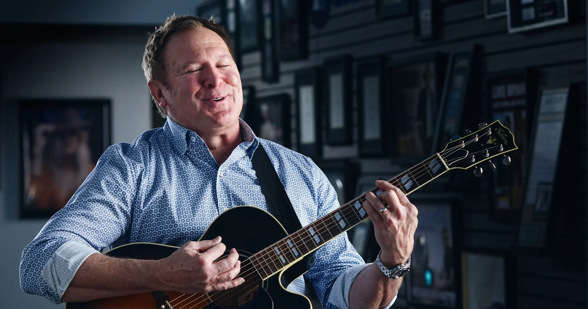 A middle-aged man in a patterned shirt playing an acoustic guitar and smiling with his eyes closed in a room decorated with framed pictures.