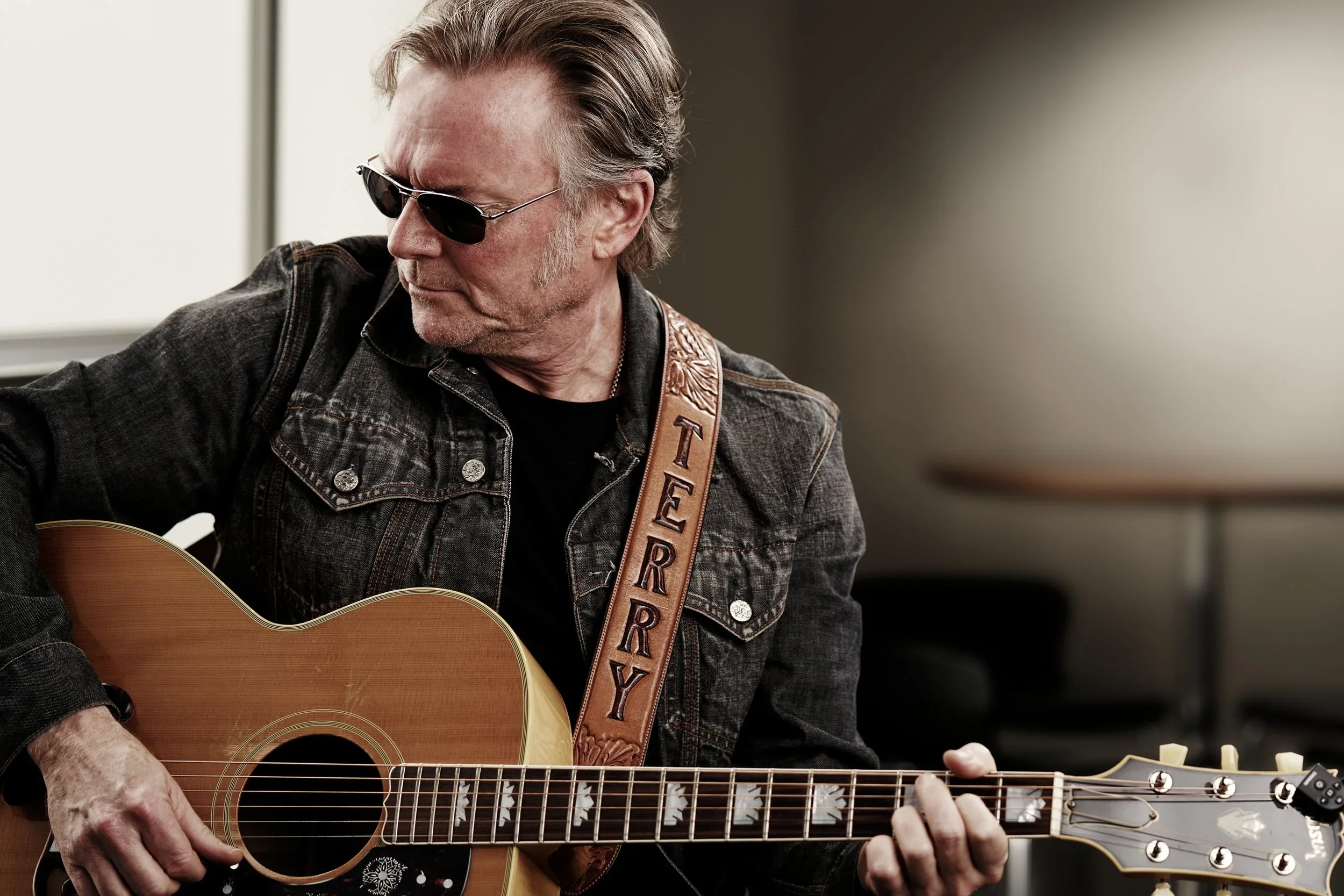 A man with gray hair and sunglasses playing an acoustic guitar. He is wearing a black denim jacket and a leather guitar strap with the name 'Terry' on it.