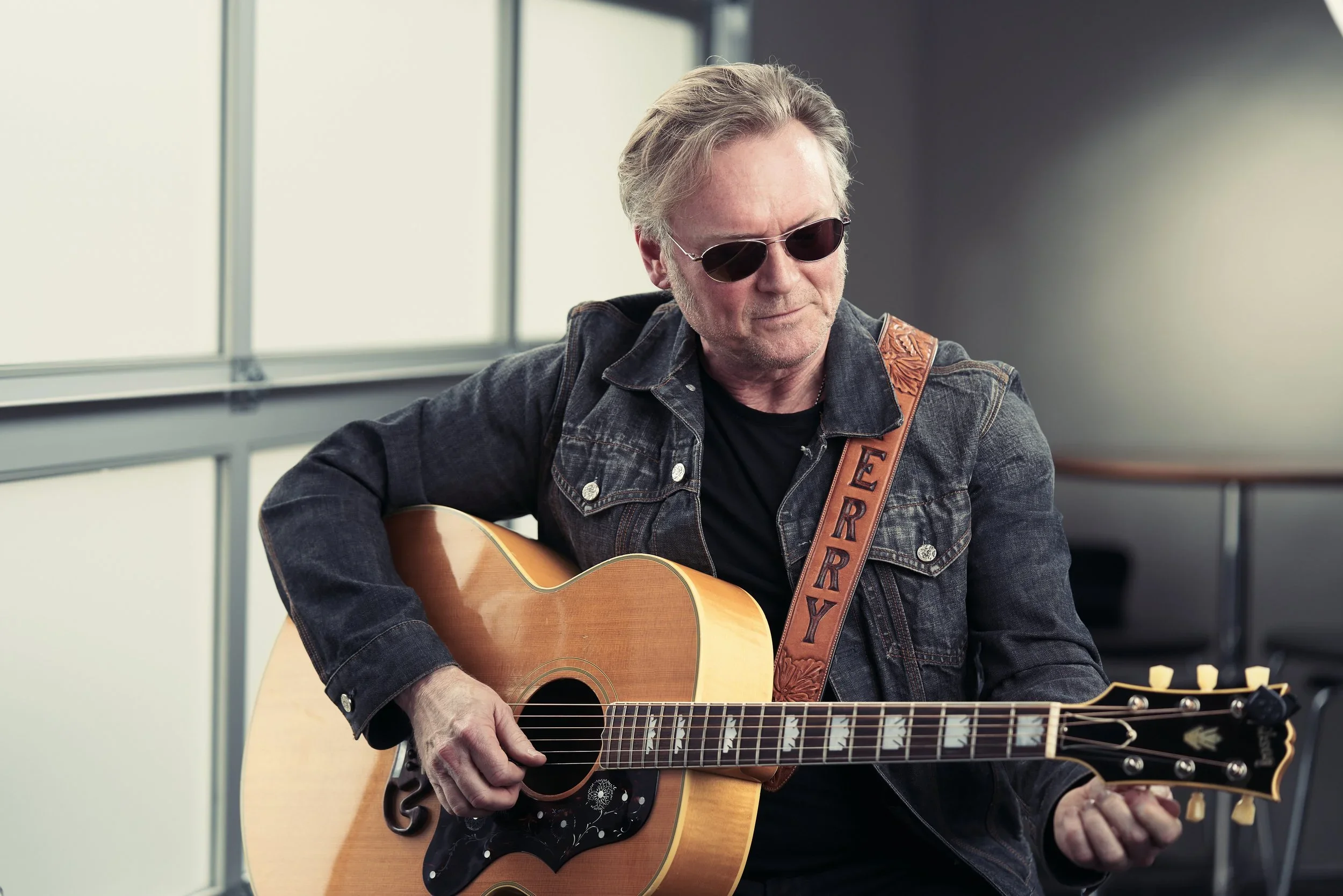A man with gray hair wearing sunglasses, a denim jacket, and a black t-shirt, playing an acoustic guitar with a decorative strap, inside a building with large windows.
