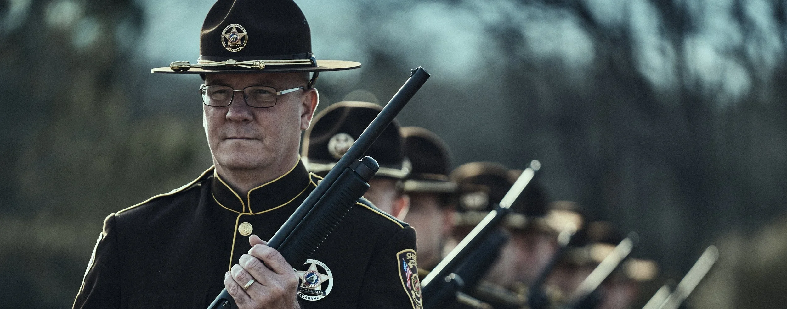 A police officer in uniform holding a shotgun with other officers in the background.