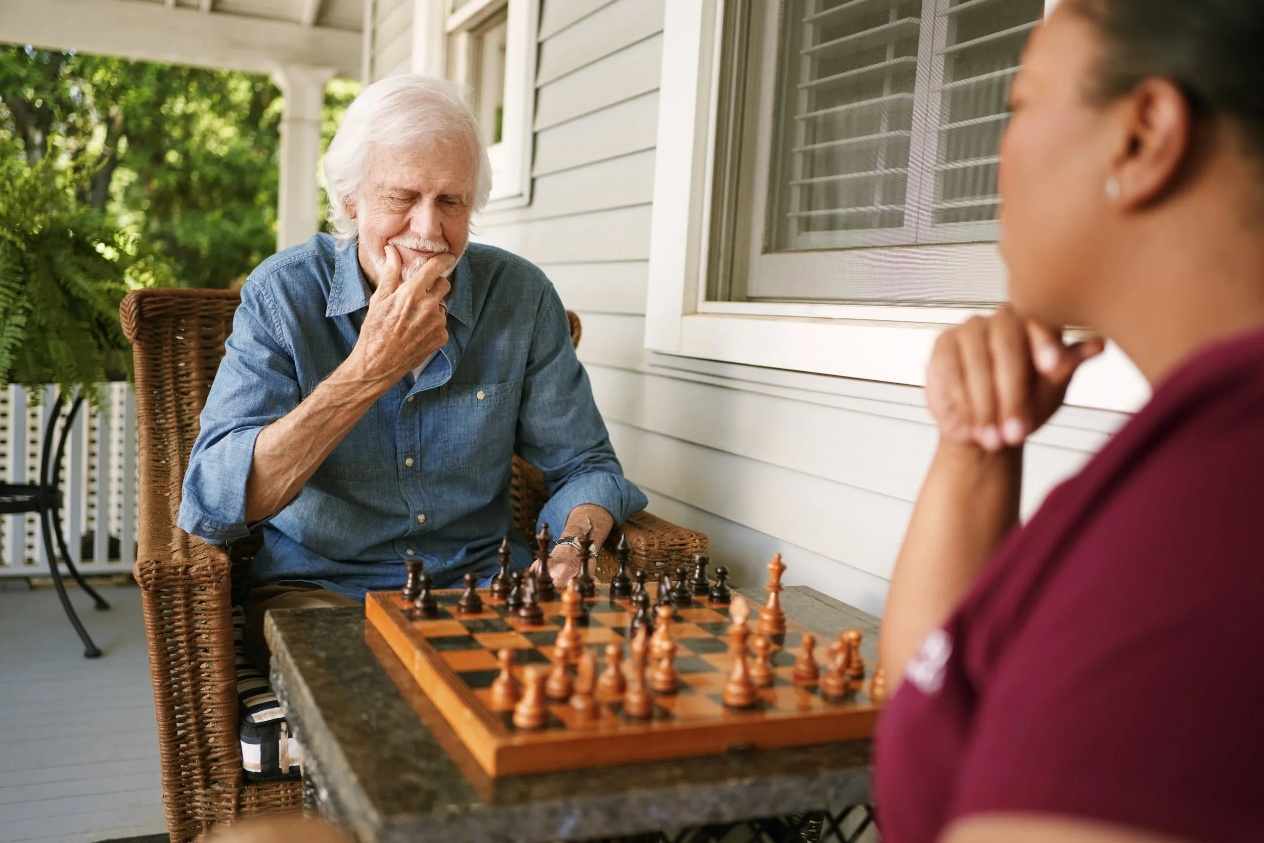An elderly man and a young woman are playing chess on a porch with a white wall and window. The man is thinking with his hand on his chin, and the woman is observing the game.