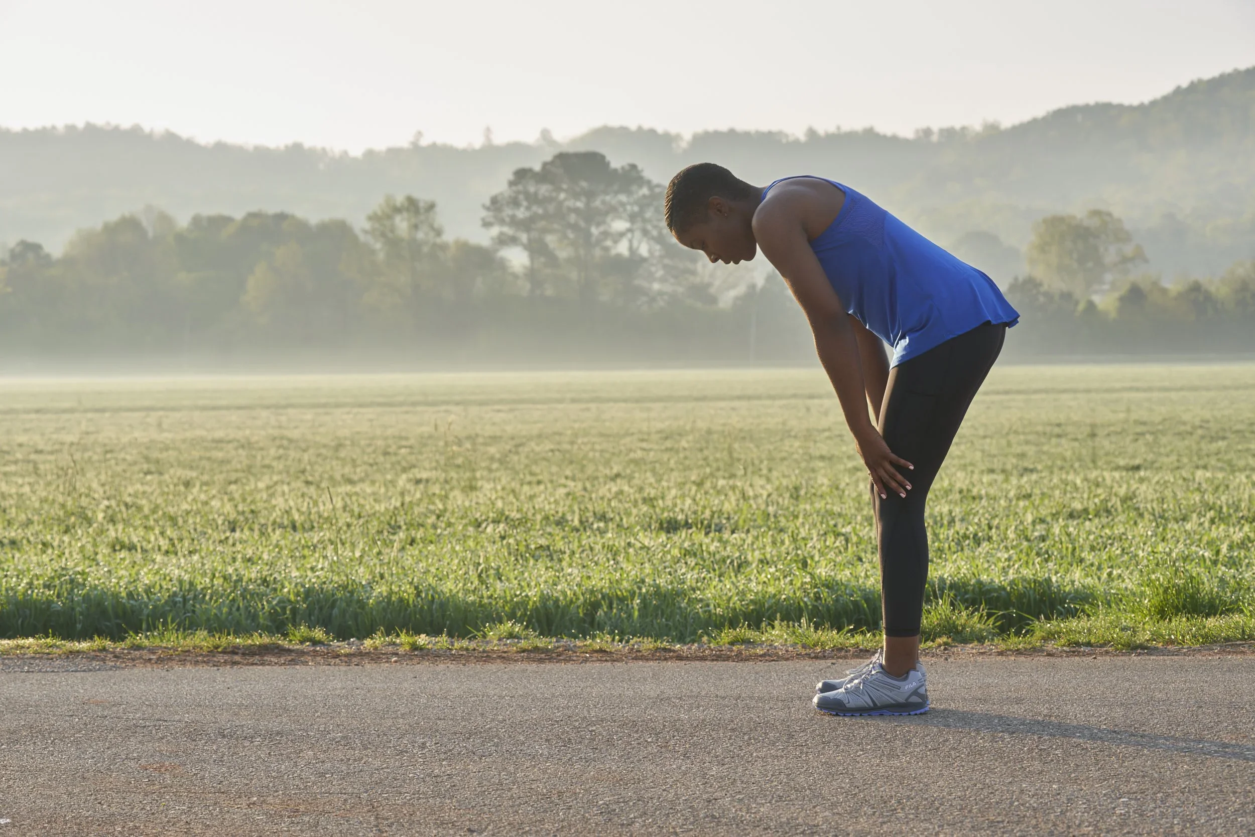 Young man in athletic attire bending over on a track, stretching in an outdoor park with trees and hills in the background during early morning.