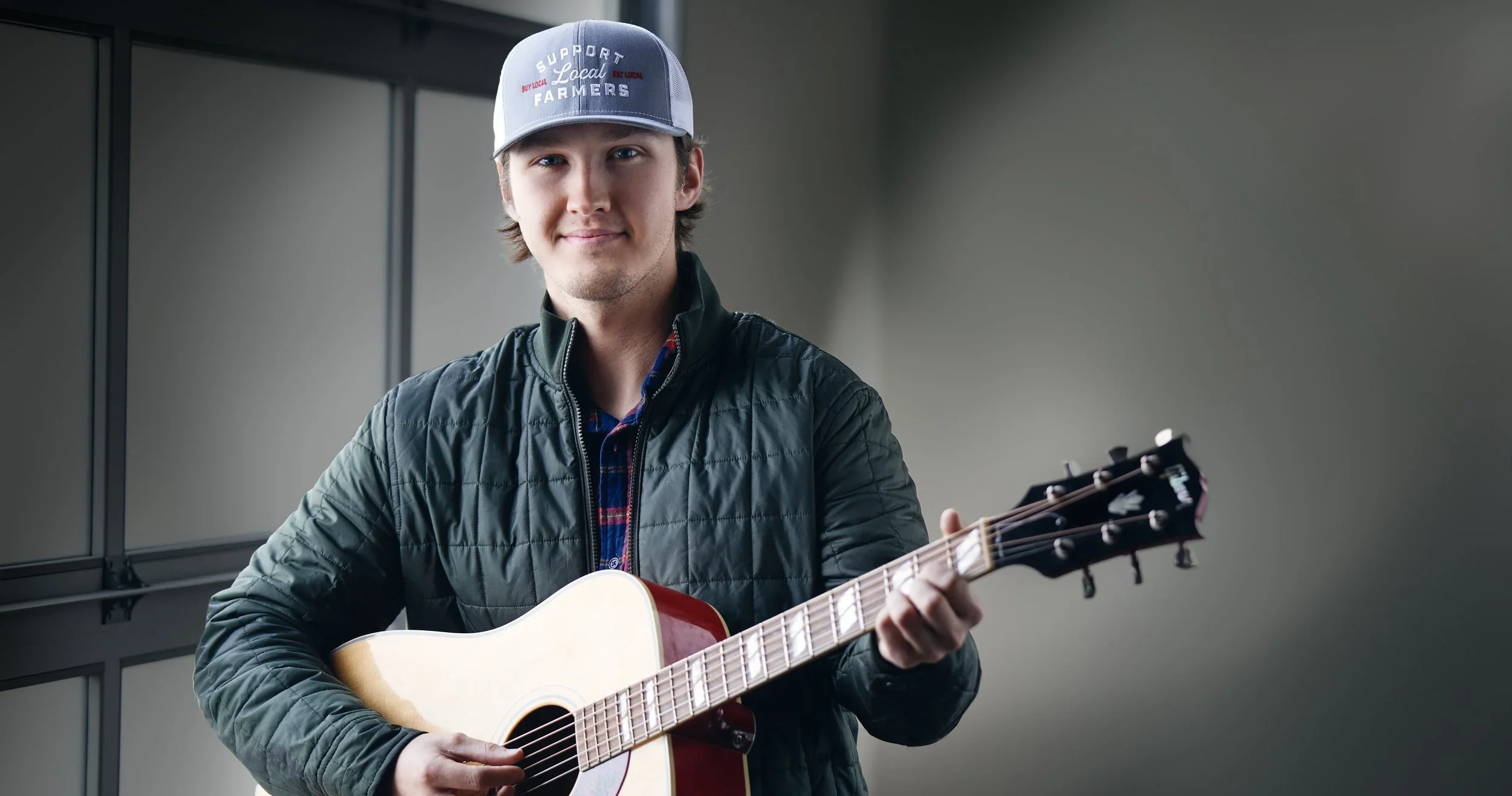 A young man smiling while playing an acoustic guitar indoors, wearing a gray cap and a black quilted jacket.