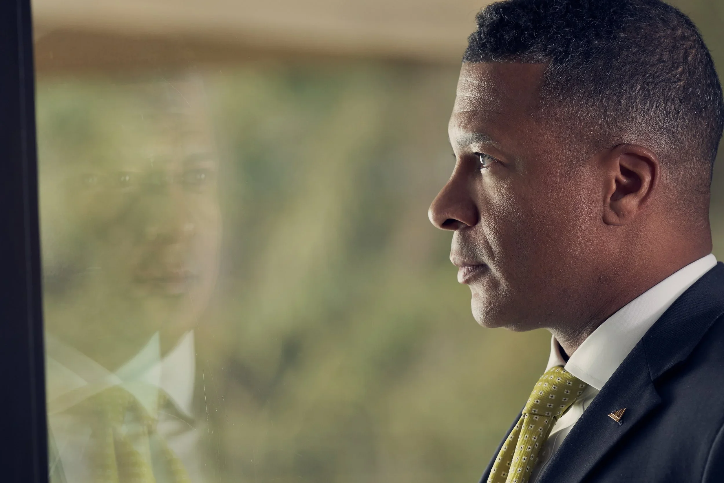 Side profile of a man in formal attire sitting and looking at a computer screen, with a reflection visible on the glass.