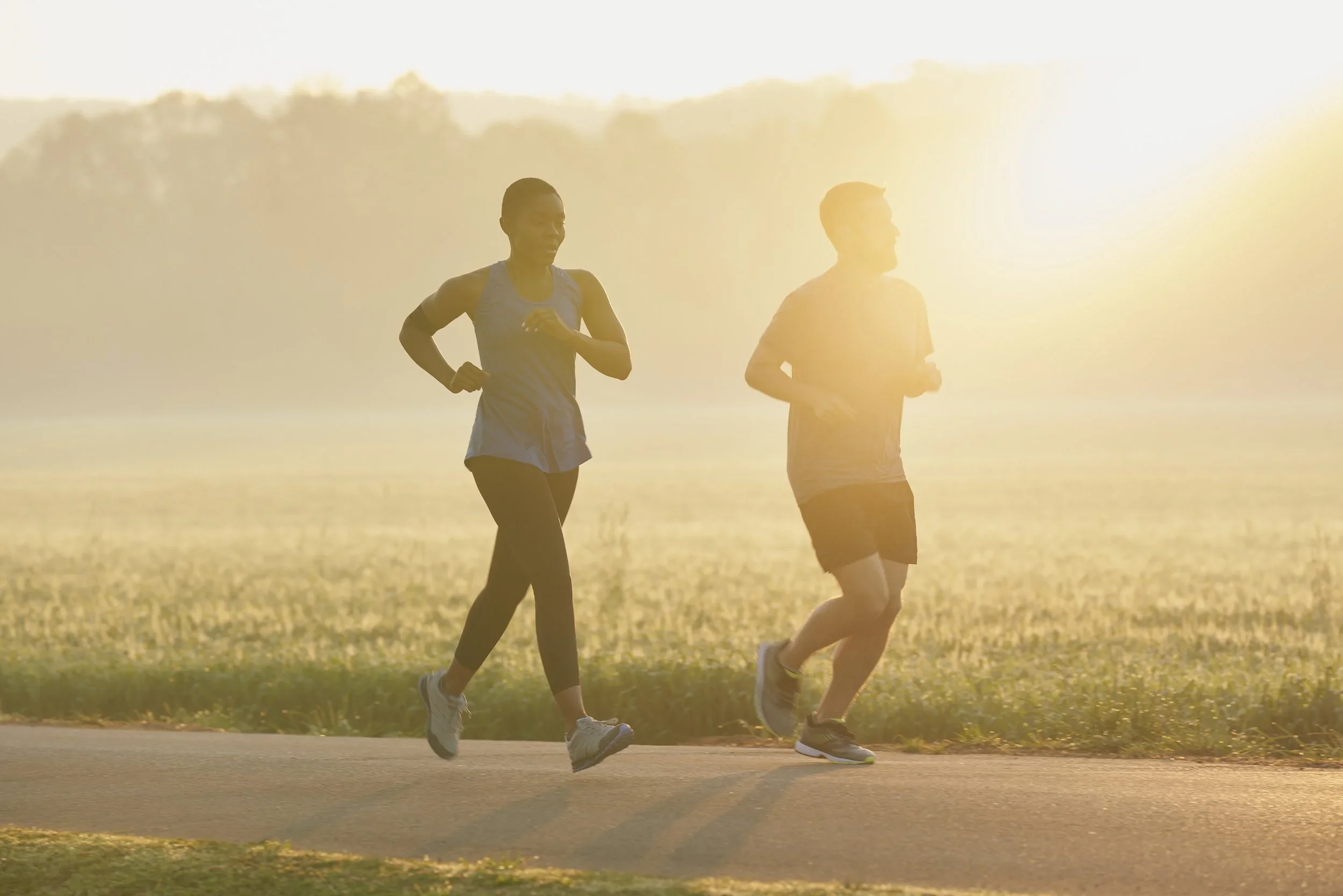Two people jogging outdoors during sunrise or sunset, with a forested area in the background and sunlight shining brightly.