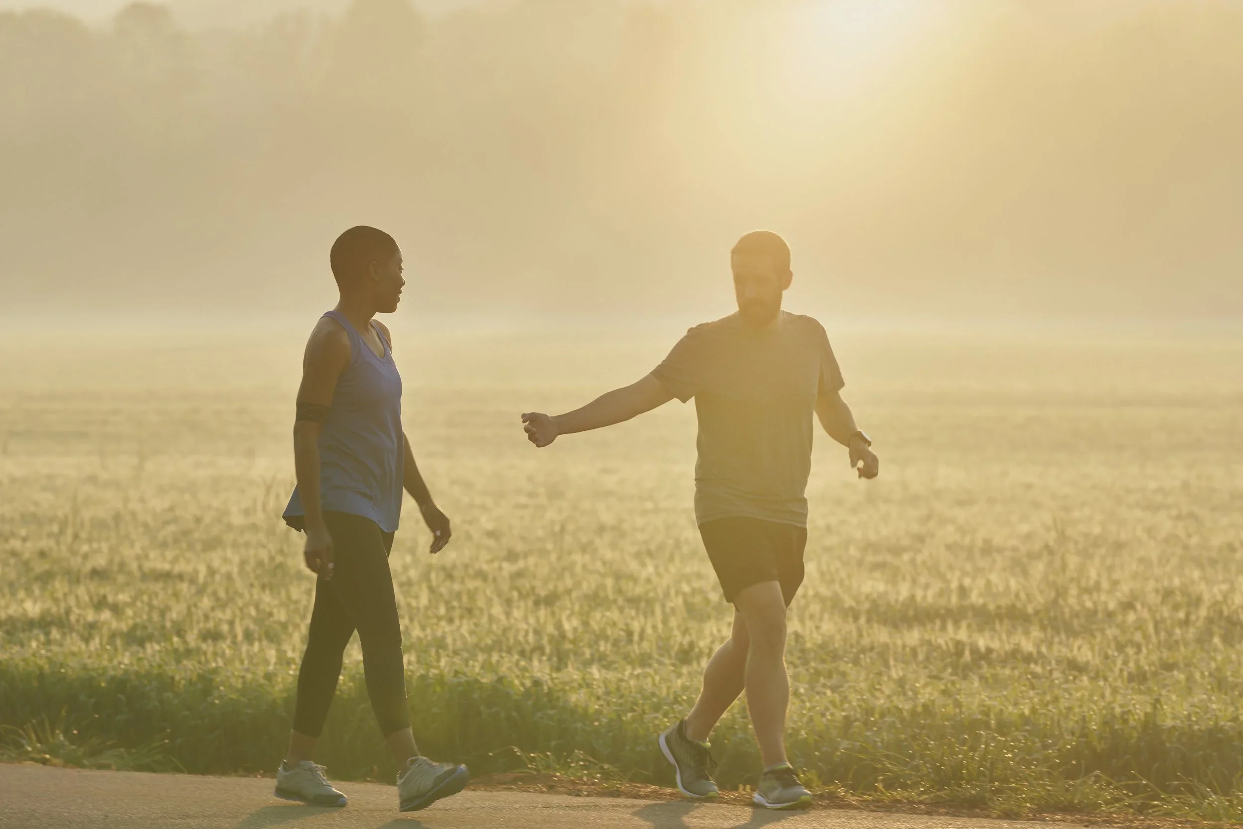 Two people walking on a path near a body of water during sunset, with one person extending their arm toward the other.