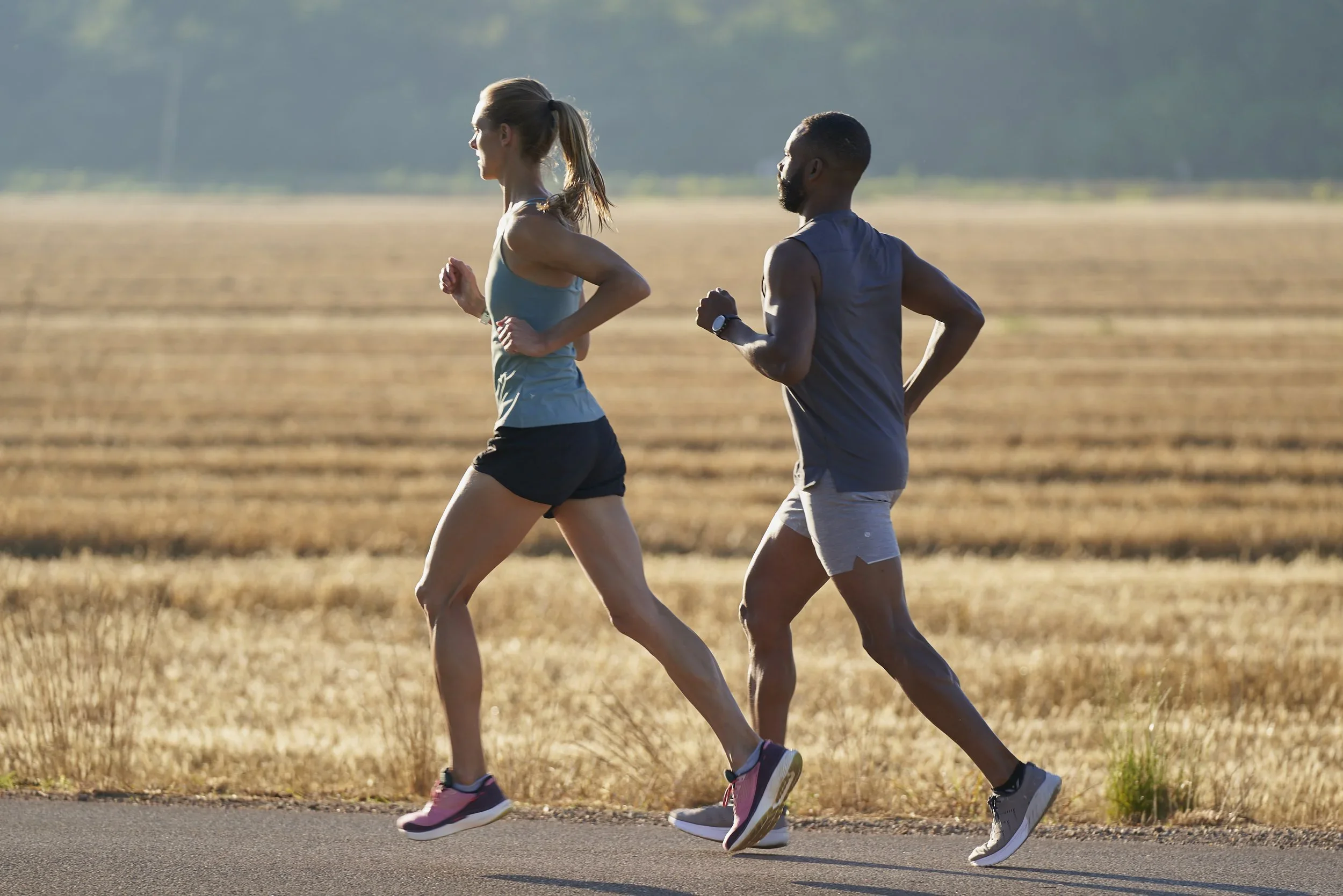 Two people jogging outdoors on a dirt path in a field, side by side, with a background of trees and clear sky.