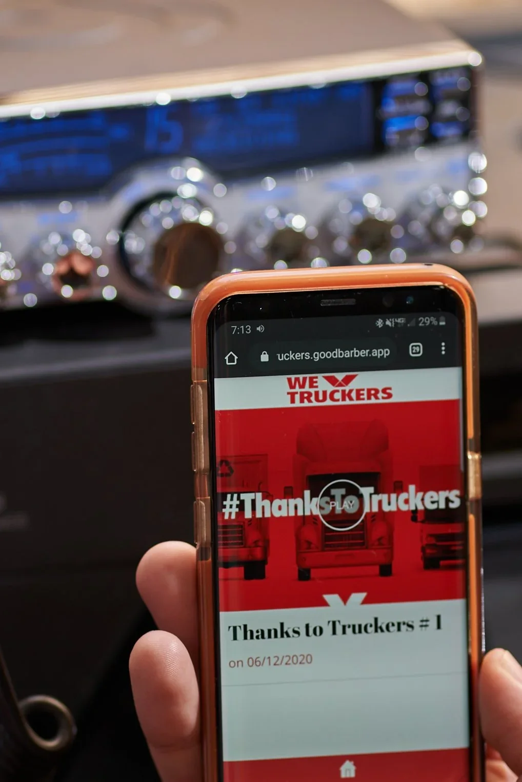 Close-up of a person's hand holding a smartphone displaying a webpage with the logo 'WE TRUCKERS' and the hashtag '#ThanksTrucker'. In the background, part of a truck or vehicle's dashboard is visible.