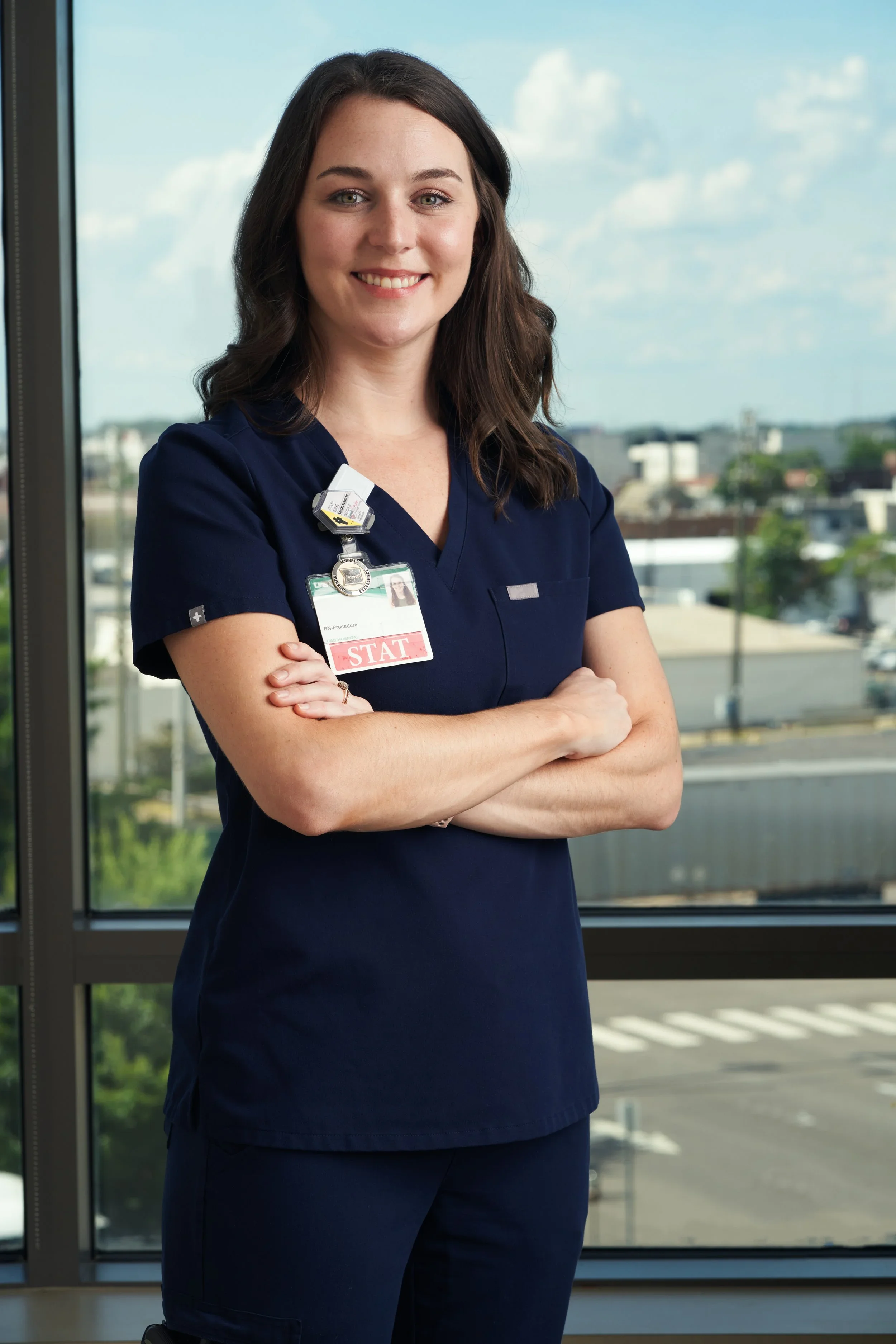 A woman in navy scrubs smiling, standing with arms crossed in front of a window with a view of a cityscape and blue sky.