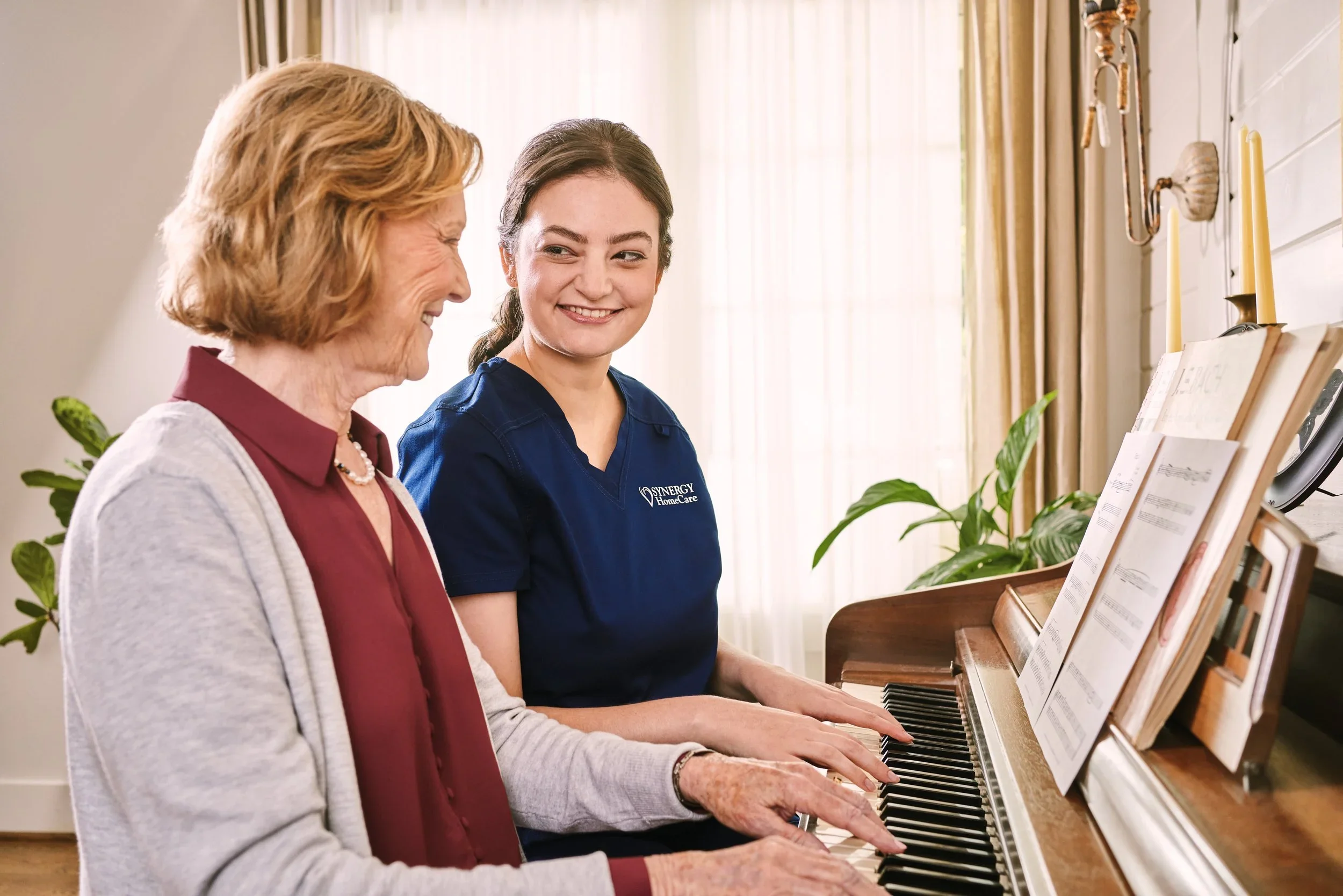 An elderly woman and a young woman smiling as they play the piano together in a bright room with curtains and plants.