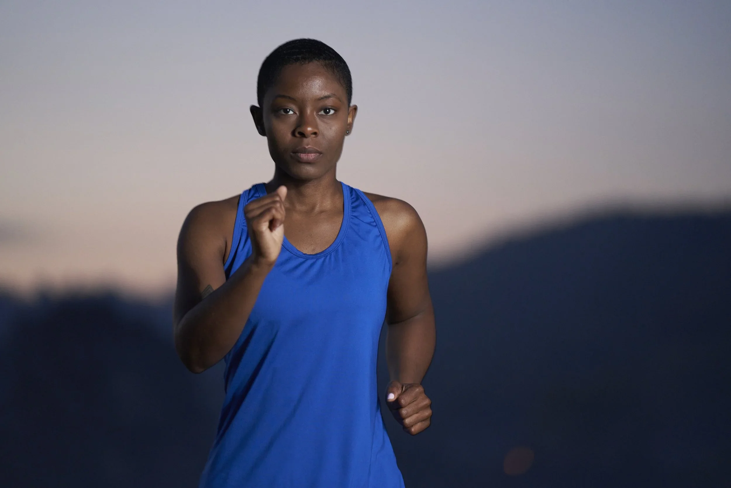A young Black woman with a short haircut, wearing a blue sleeveless athletic shirt, running outdoors during dusk or dawn.