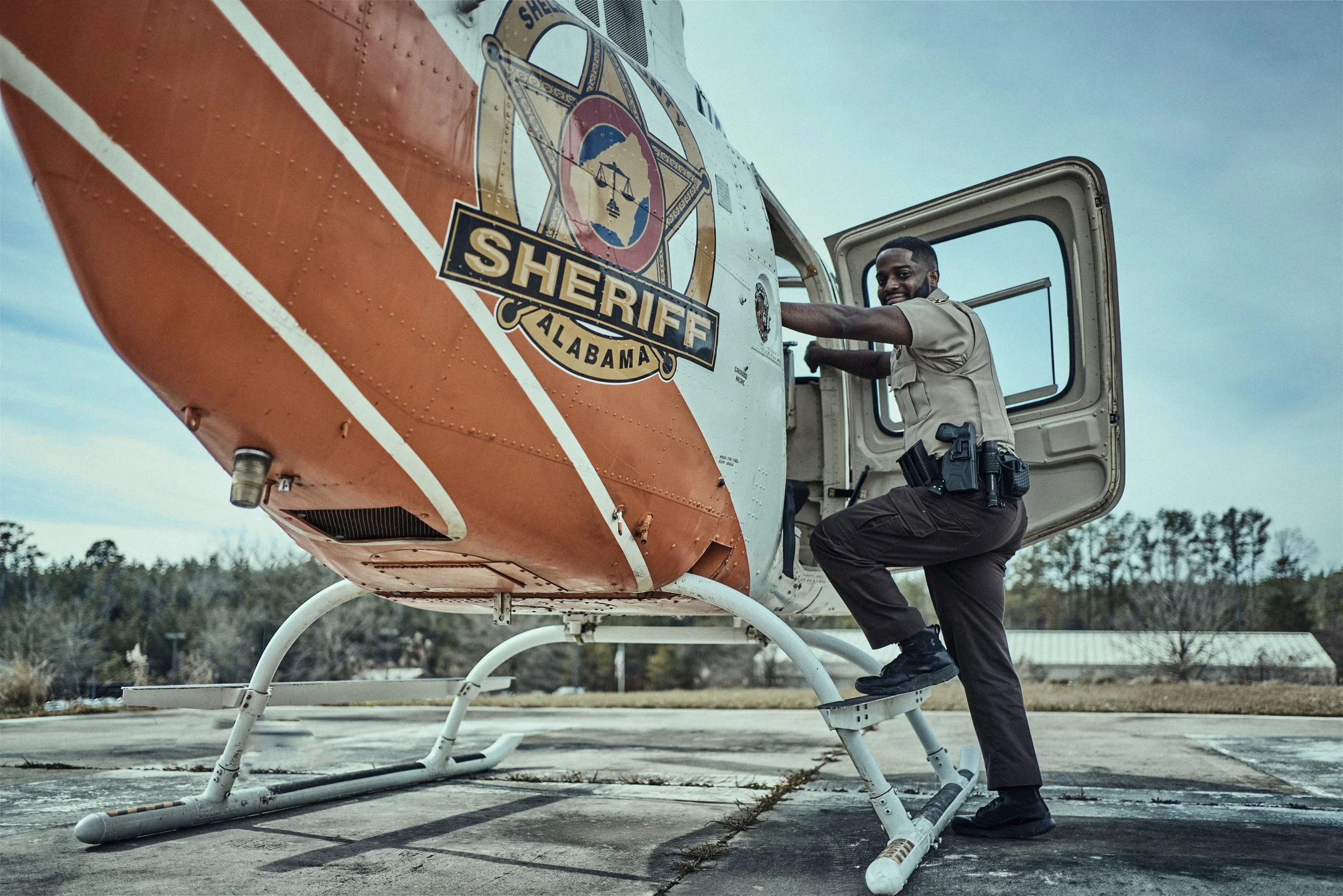 A police officer in uniform is kneeling and leaning out of a helicopter's open door, looking at the camera. The helicopter is marked with a sheriff's badge from Alabama.
