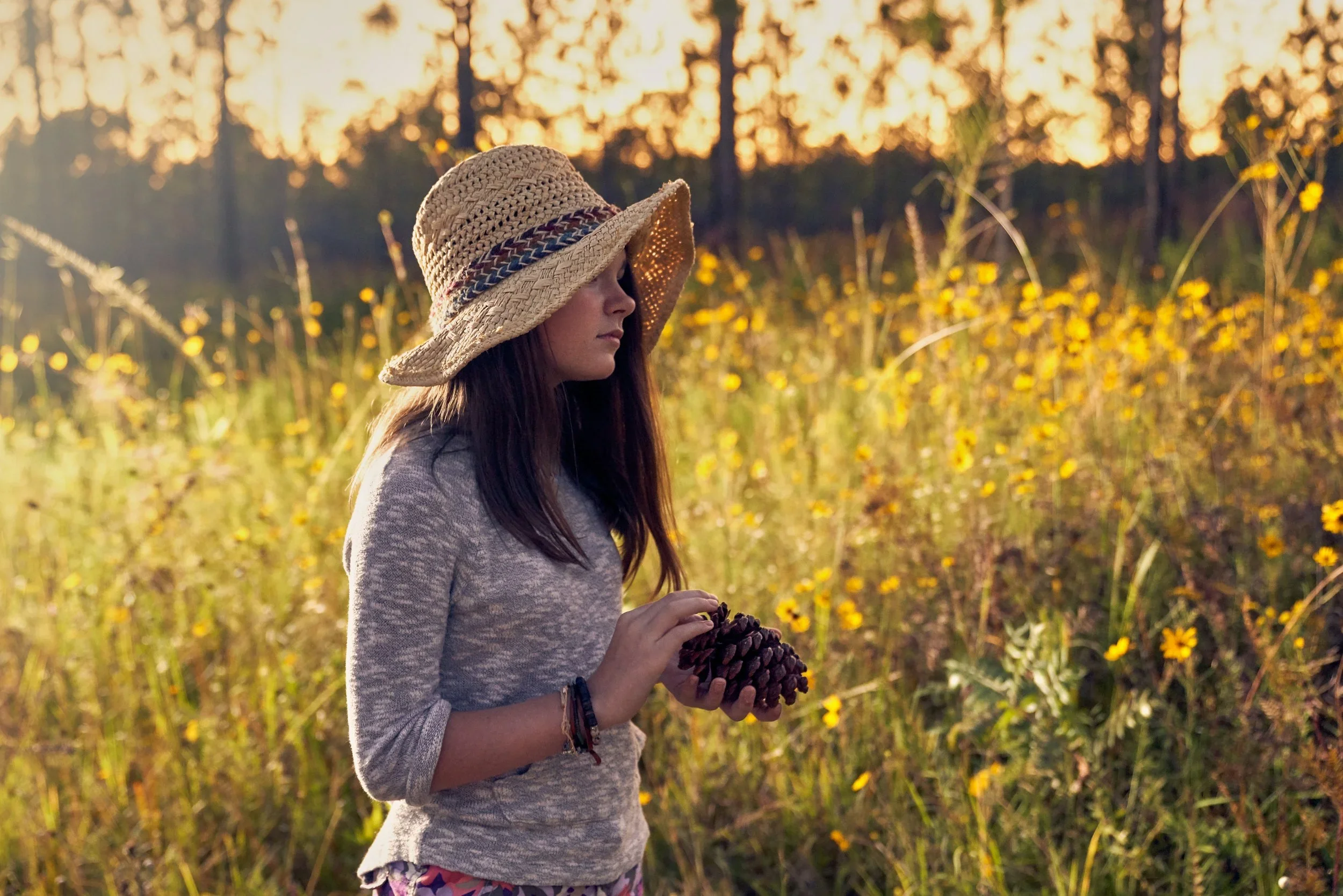 A woman in a straw hat with a woven band, wearing a long-sleeve gray shirt, standing in a field of yellow flowers during sunset, holding a large pine cone.